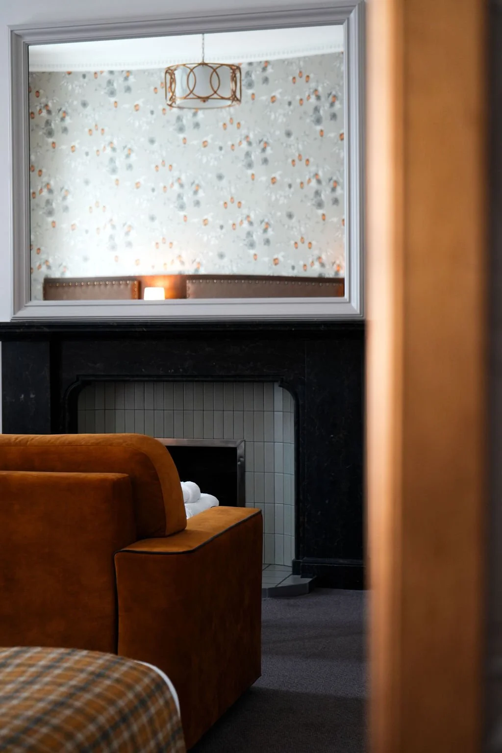 A cozy living room with a brown velvet armchair, a fireplace with light gray tiles and a dark surround, a mirror reflecting a decorated wall, a pendant light hanging from the ceiling, and a small lit lamp on the wall.