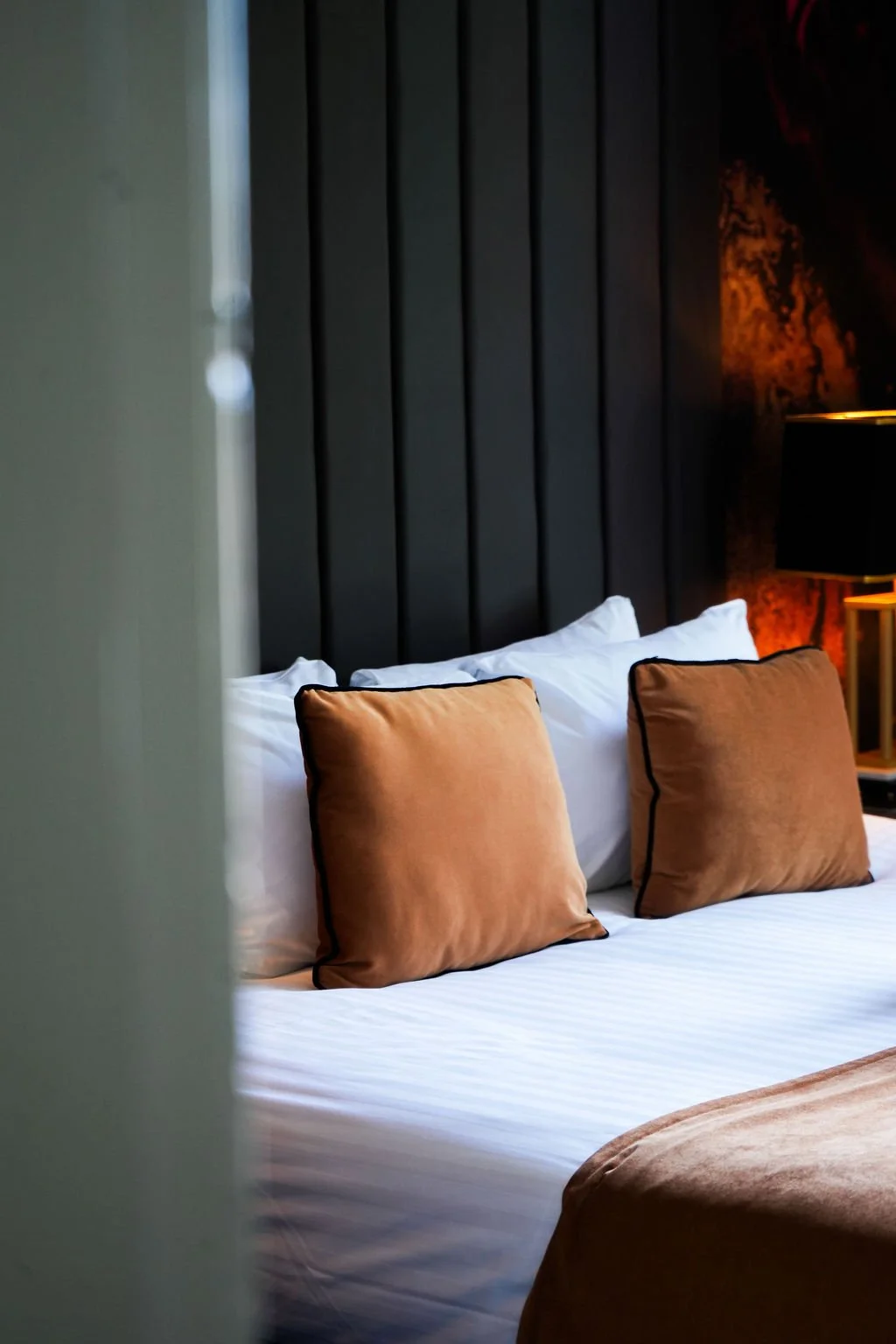 A neatly made bed with four white pillows against a dark wooden headboard in a hotel room, as seen through a partially opened door.