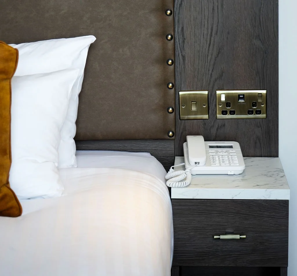 Close-up of a bedroom nightstand with a white telephone, set against a dark wood headboard with metal accents and electrical outlets.