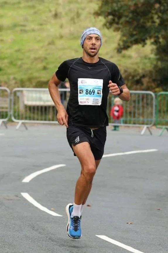 Man running in a race, wearing a black T-shirt and shorts, with a blue headband, race bib number 869, and blue running shoes.