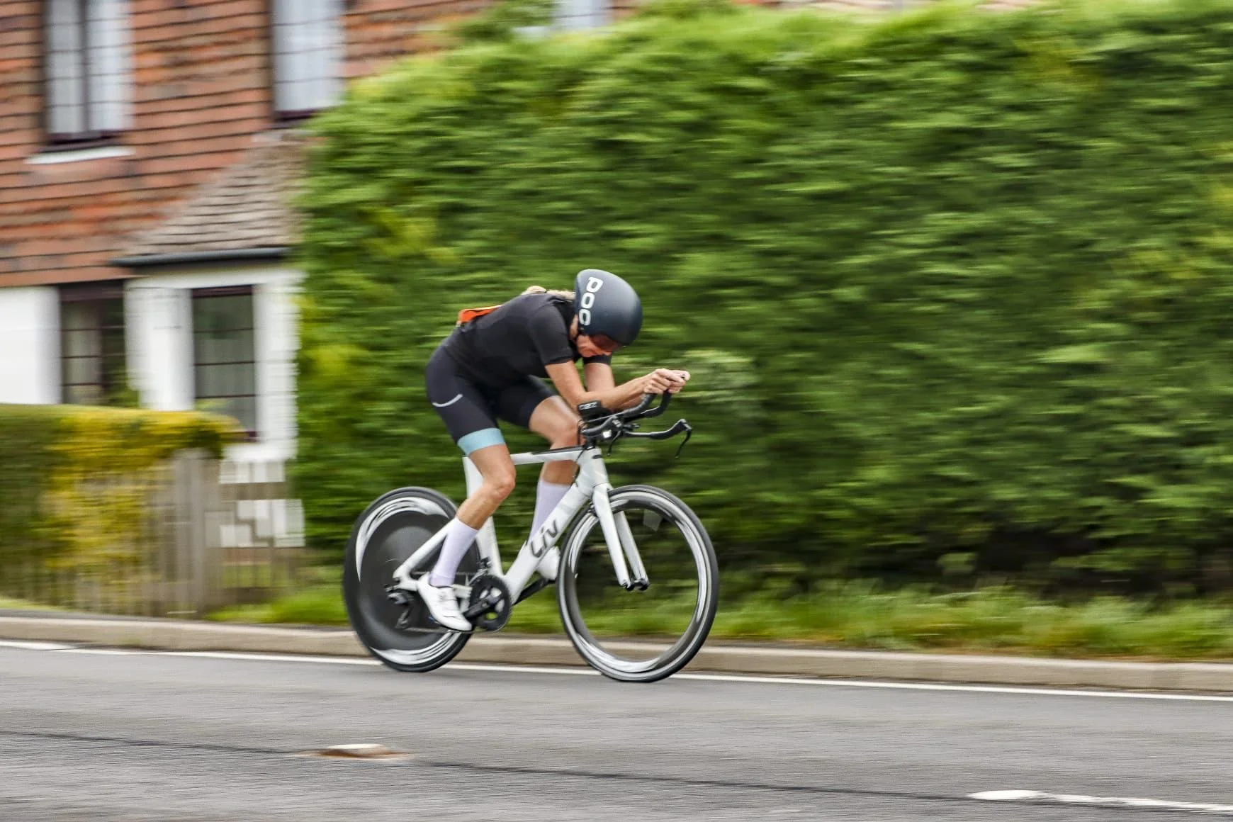 A person wearing a black helmet, black clothing, and white cycling shoes riding a white road bike on a residential street, with a green bush and a brick house in the background.