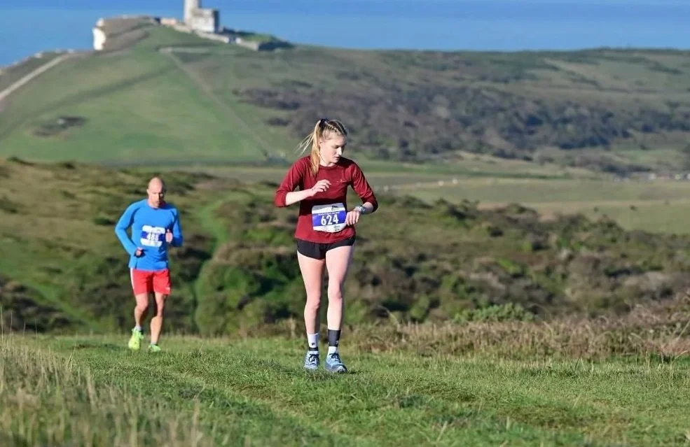 Two runners participating in a race on a grassy trail outdoors, with a hill and a building in the background.