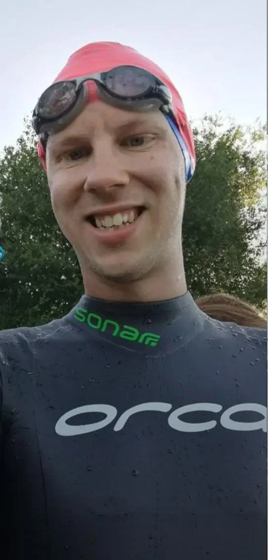 Close-up of a smiling young man wearing a red swim cap, black goggles on his forehead, and a black wetsuit with green and white branding, outdoors with trees in the background.