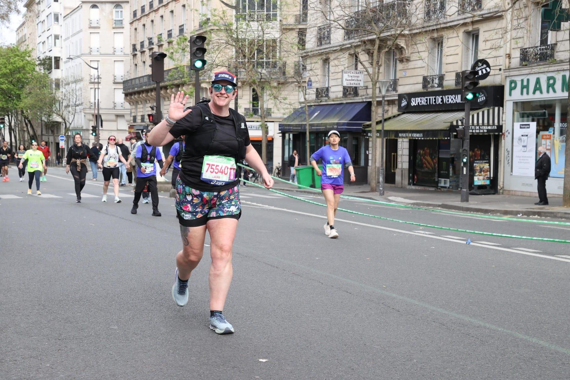 A woman runs a marathon in the city, waving and smiling at the camera, with other runners behind her and city buildings on the street.