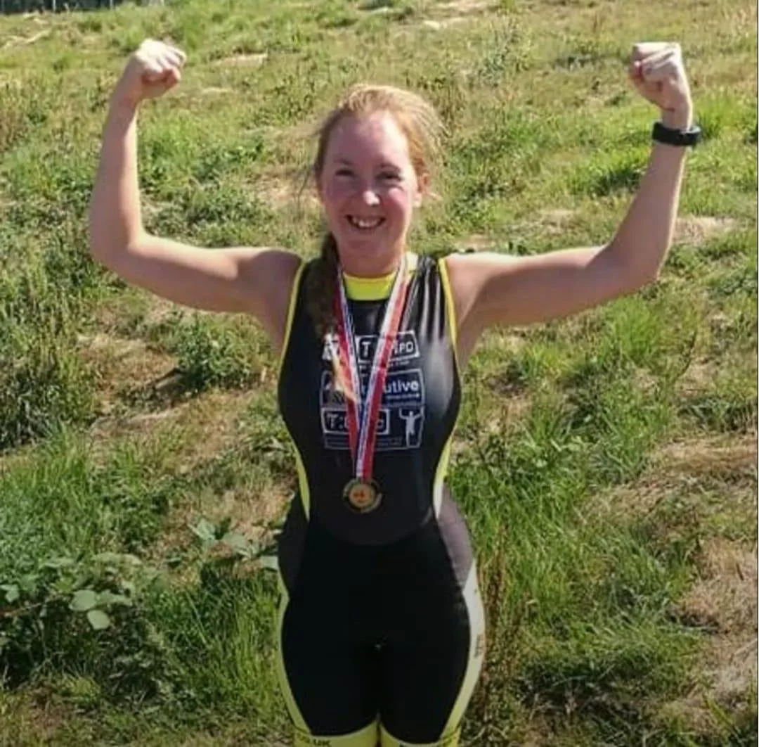 Young woman in athletic gear, smiling and flexing her biceps, wearing a medal around her neck, standing outdoors on grass.