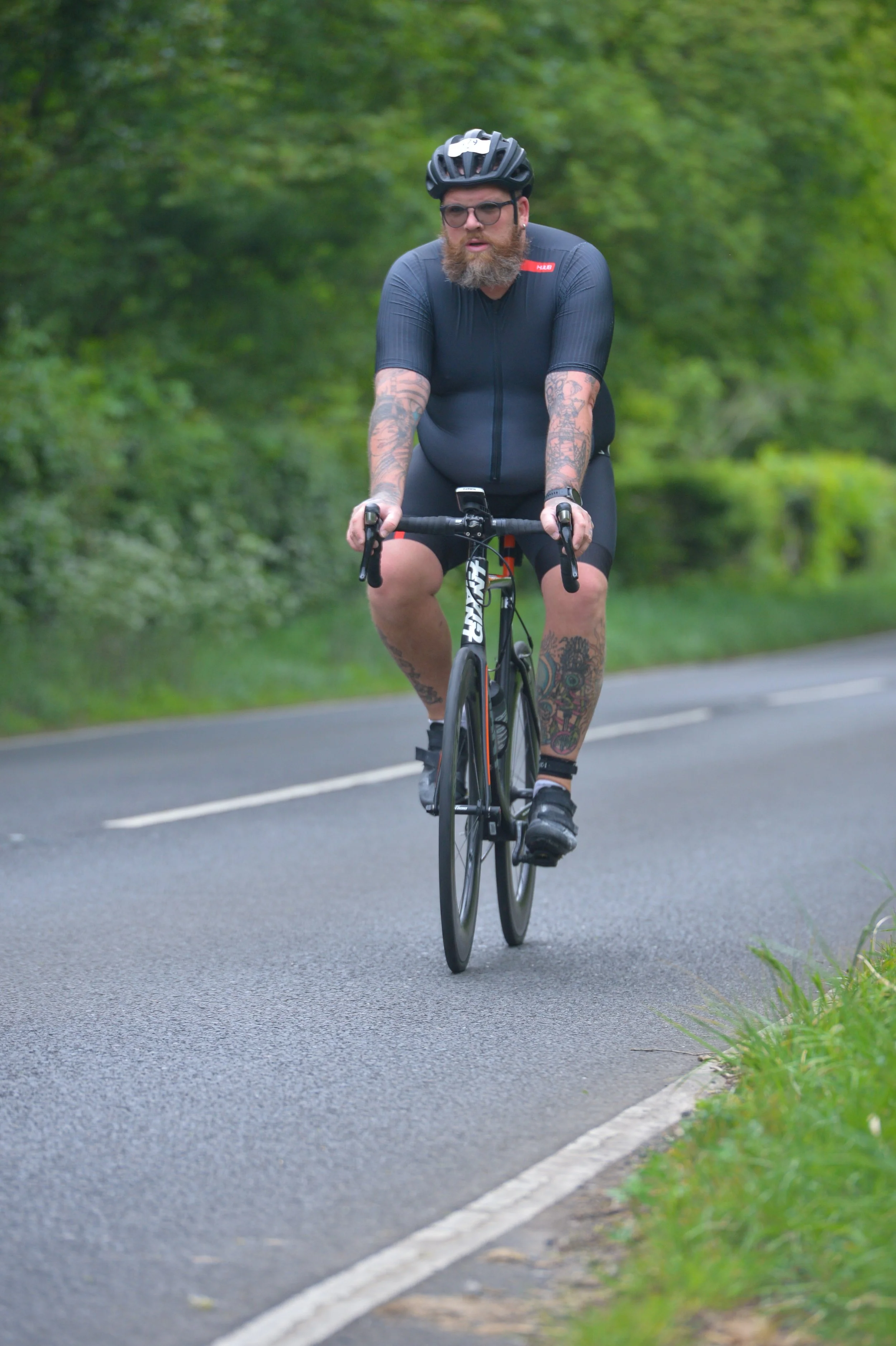 A man with a beard and tattoos riding a black road bike on a paved road surrounded by green foliage.