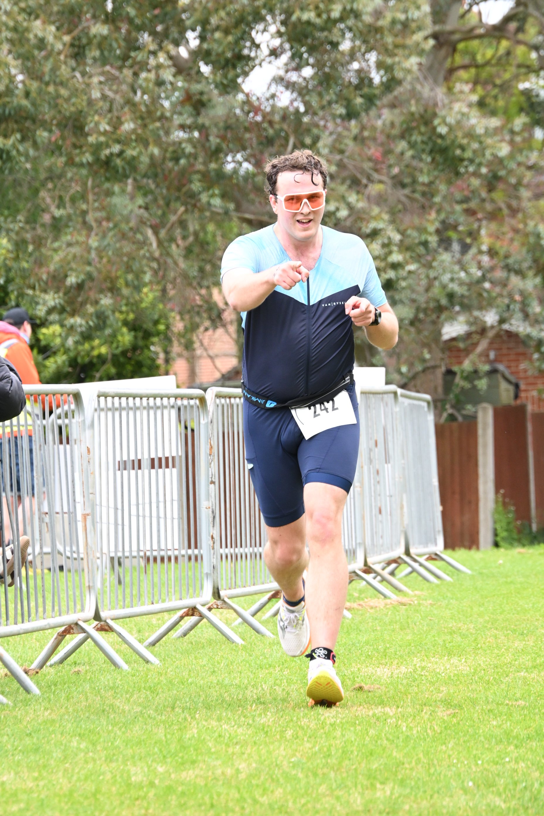 Male runner wearing sunglasses and a race bib numbered 242, running outdoors on grass, near a white metal fence during a race event.