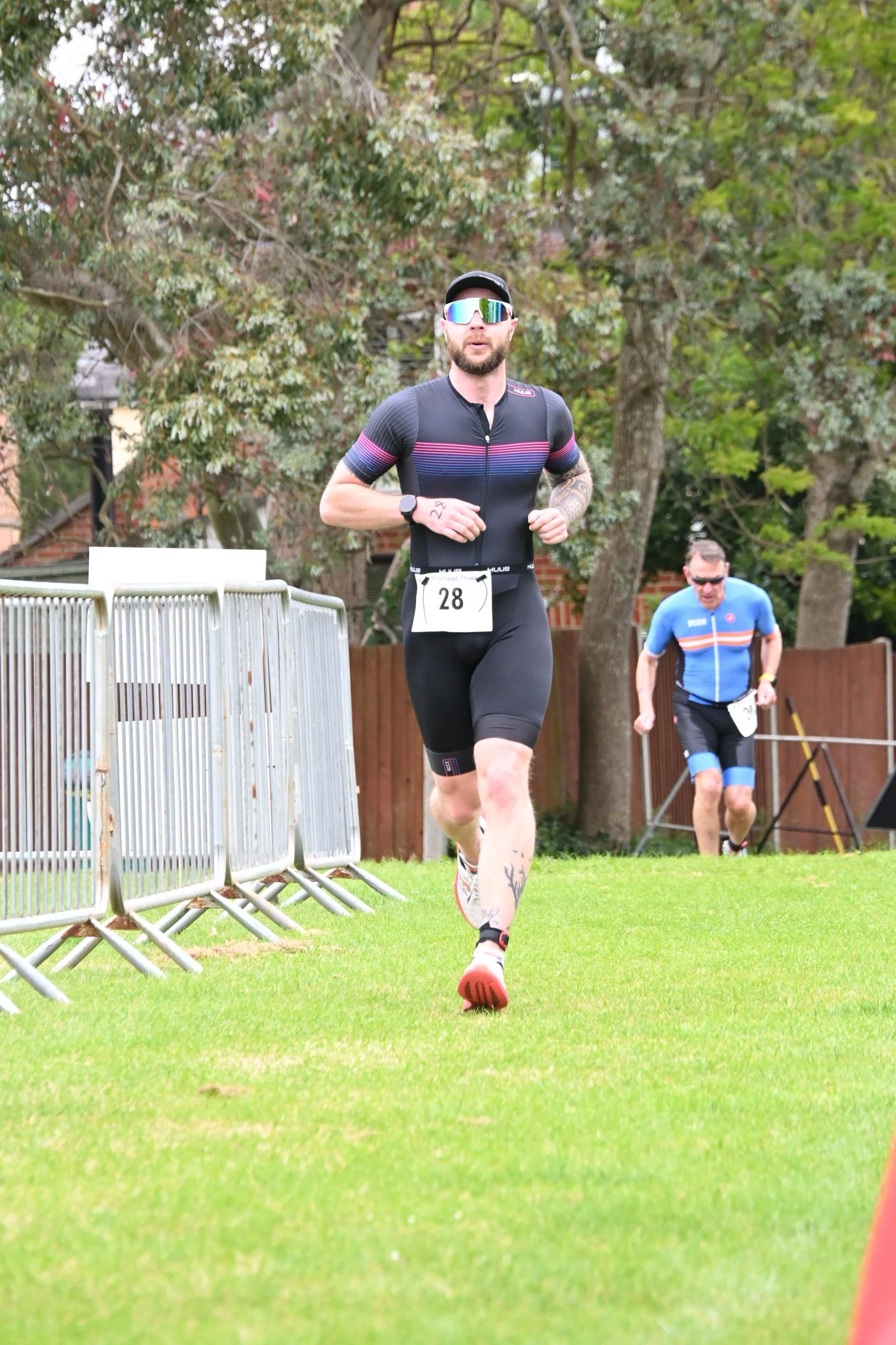 Male runner wearing sunglasses, black athletic outfit with pink and purple stripes, running on a grassy area, with a second male runner in blue in the background, surrounded by trees and a fence.
