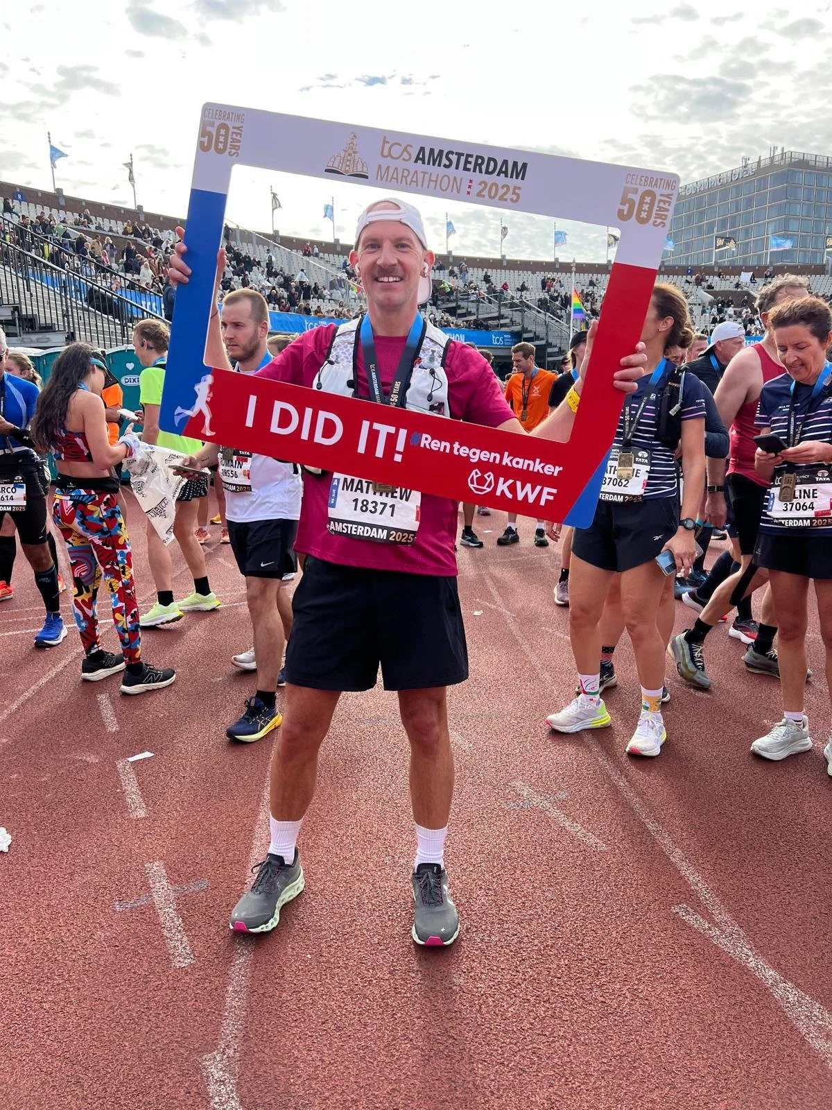 Marathon runner holding a frame with the words "I DID IT!" at a race event, with other runners in the background at a stadium in Amsterdam, 2025.