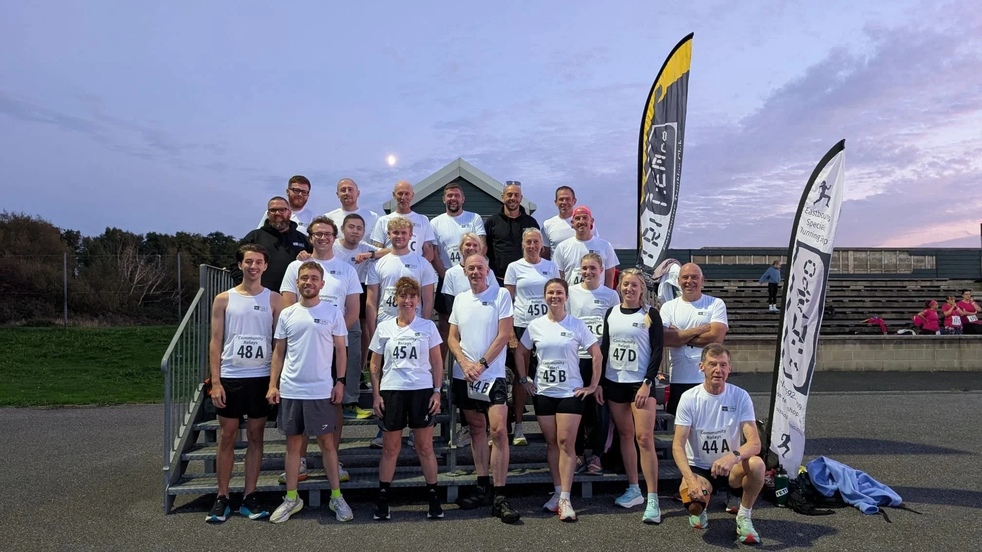 A group of people, some wearing white t-shirts with race numbers, posing on a staircase outdoors during an event in the evening, with flags and other people in the background.