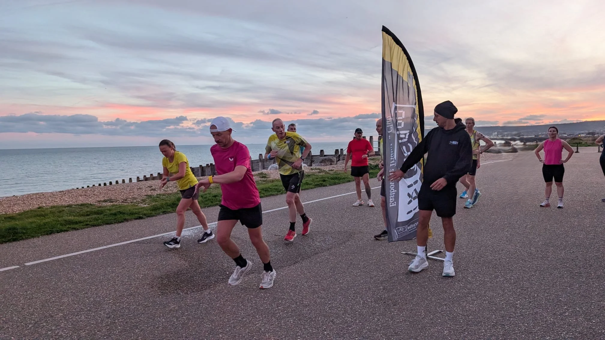 Group of runners participating in a race along a coastal path at sunset, with a flag that reads 'Explore' and a calm sea in the background.