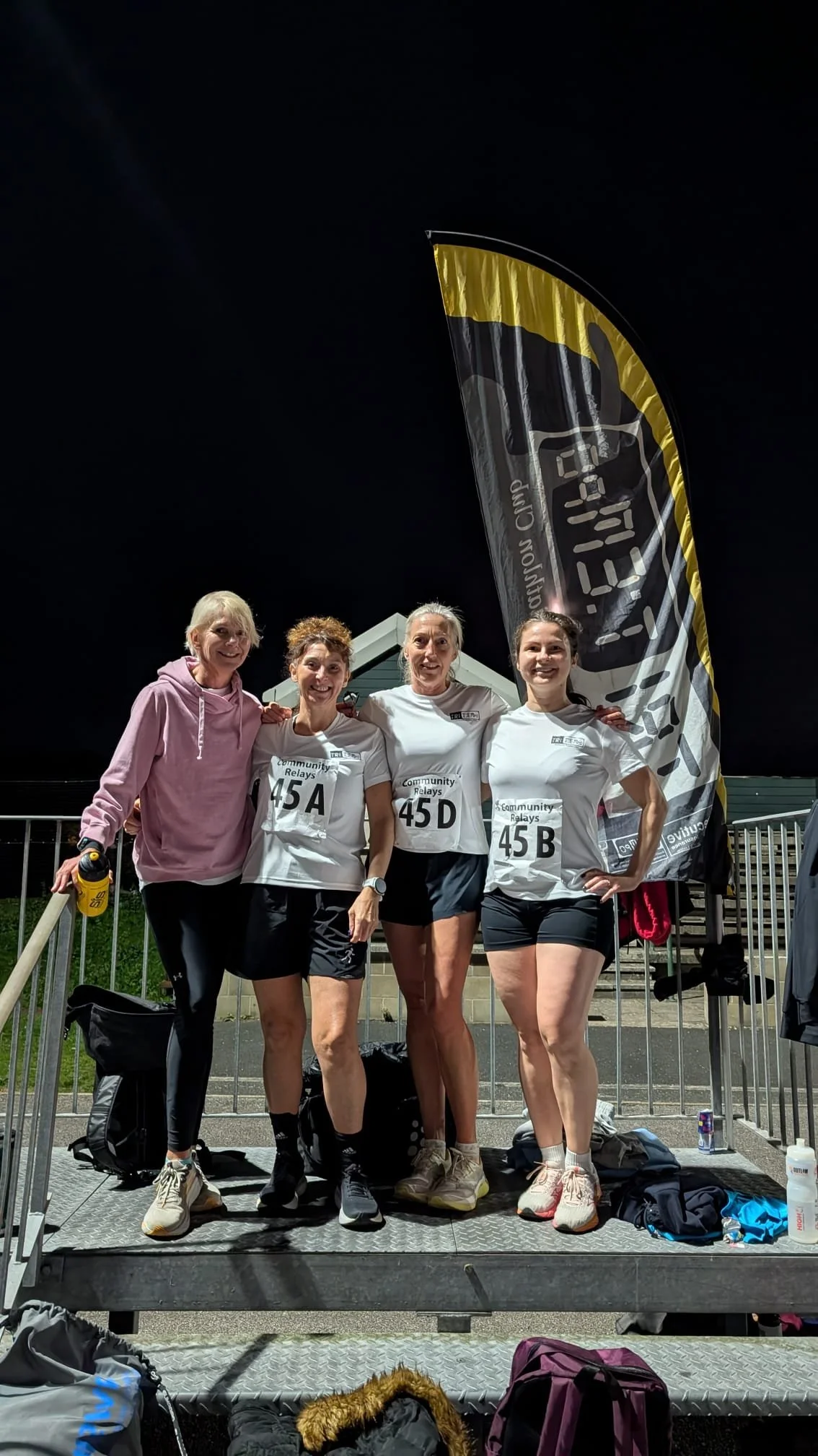 Four women in athletic gear standing together on a raised platform after a race at night, with a tall black and yellow flag behind them that reads 'Community Relays' and a dark sky overhead.