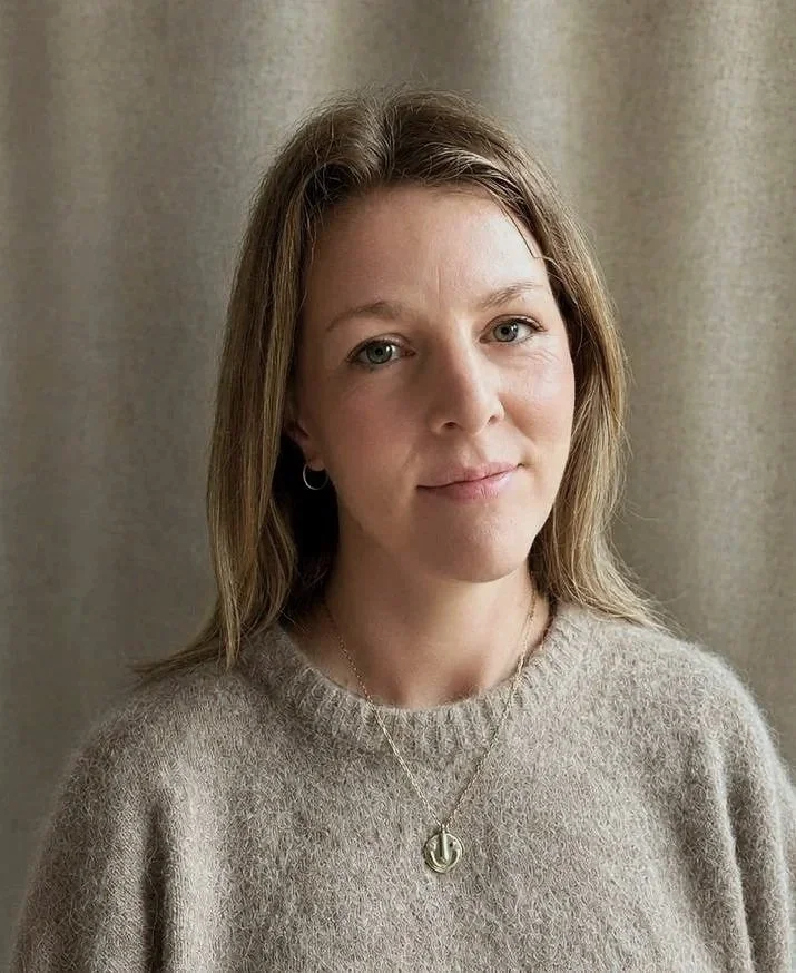 A woman with brown hair and blue eyes wearing a beige sweater, a necklace with a circular pendant, and small hoop earrings, standing indoors against a curtain background.