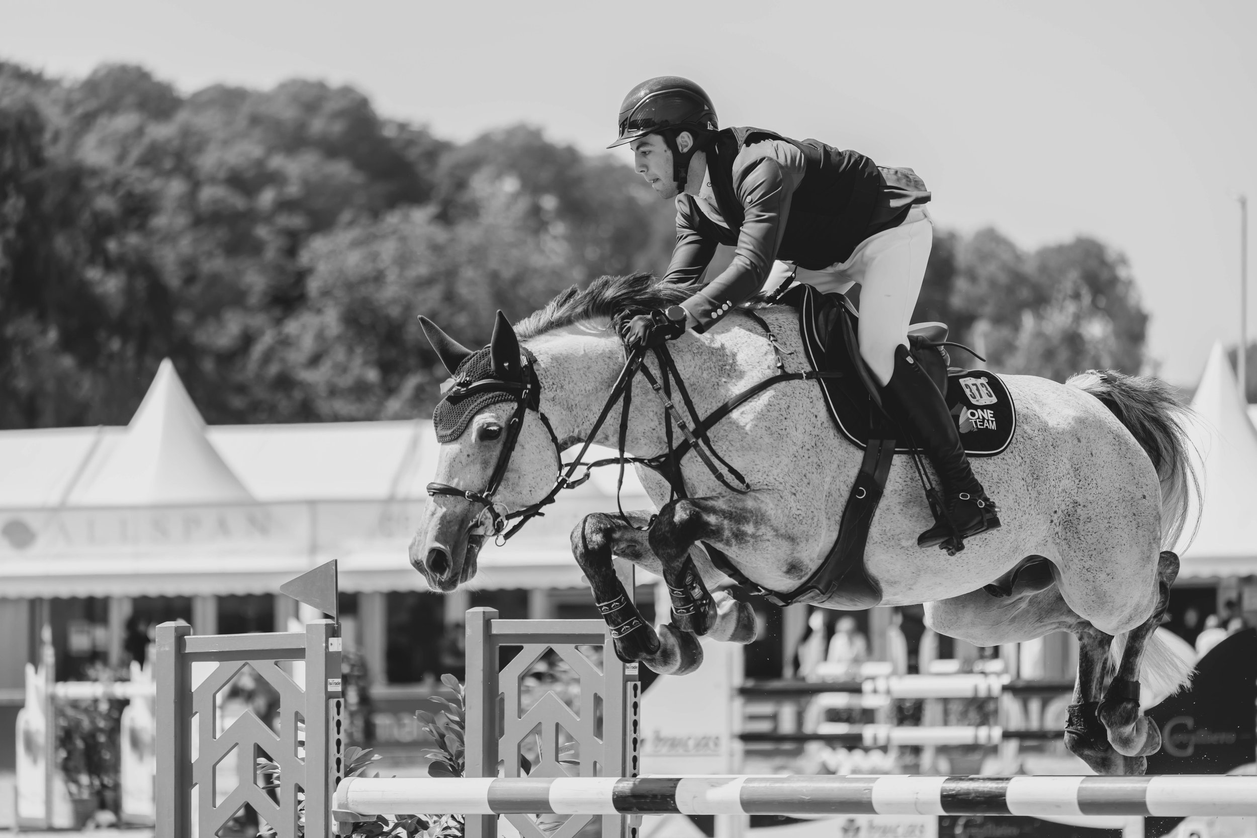 A male equestrian rider in a helmet and riding attire who is in mid-air on a horse, participating in a show jumping competition, with jumps and a tent in the background.