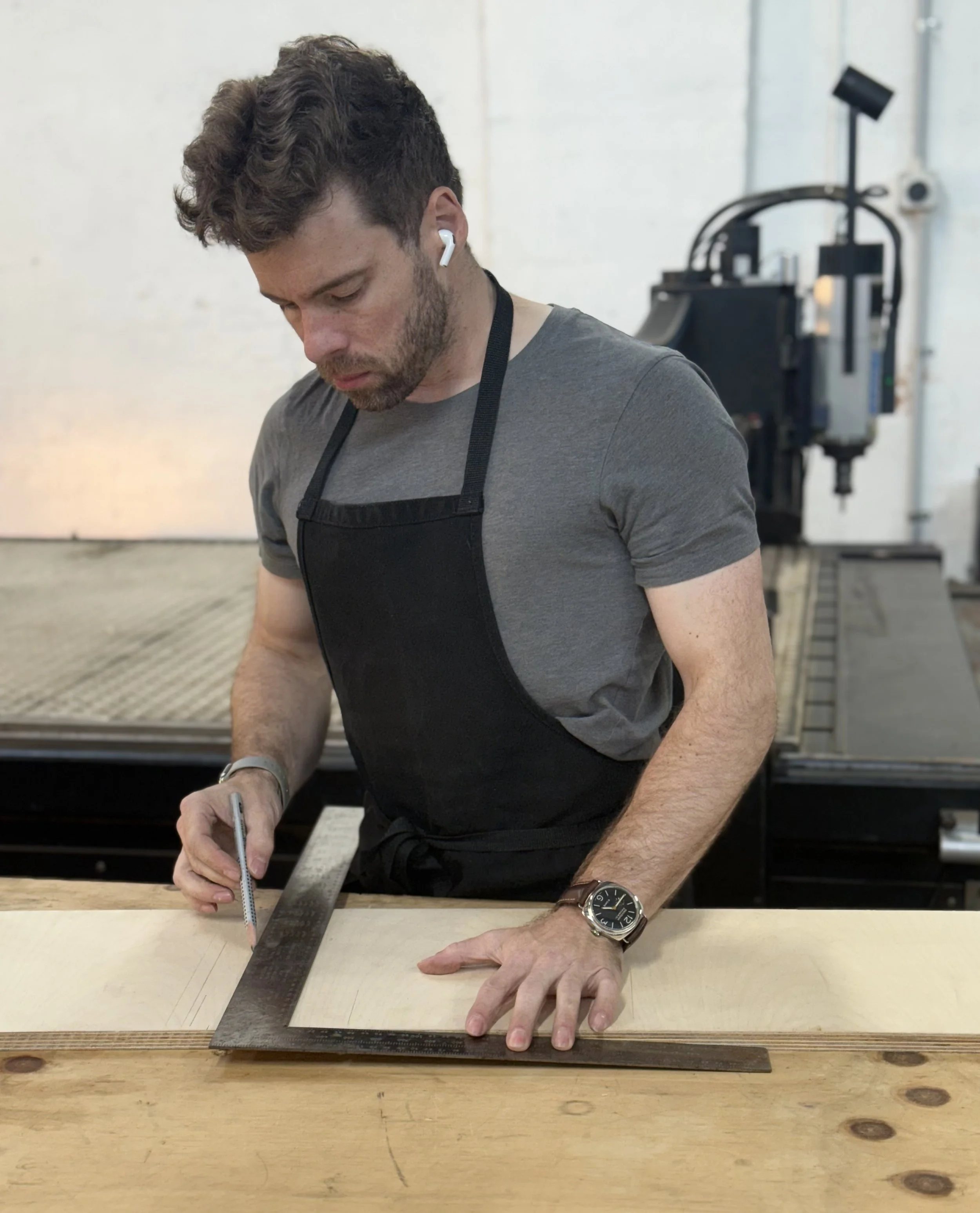 A photo of James wearing a charcoal t-shirt and a dark black denim apron. He is using a steel square to mark a piece of birch plywood with a pencil. A large industrial CNC router can be seen in the background.