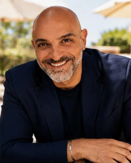 A smiling bald man with a beard wearing a dark blazer, seated outdoors with umbrellas and greenery in the background.