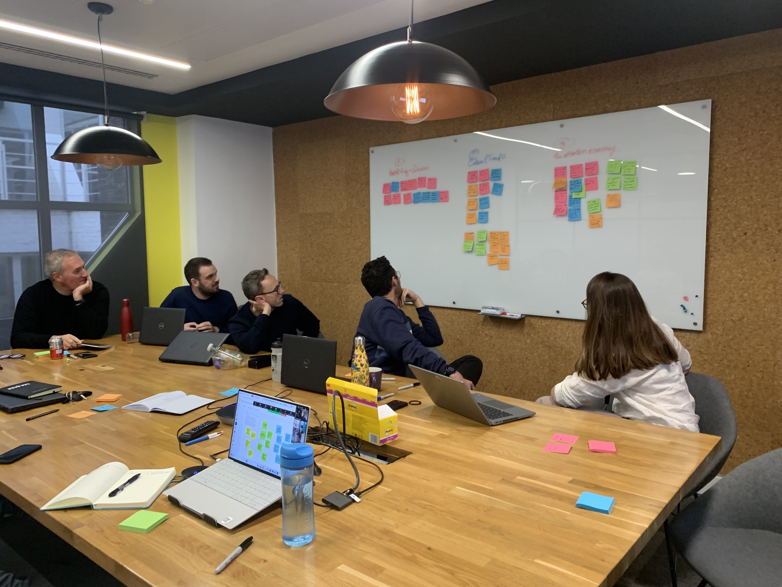 A group of people seated around a large wooden table in a meeting room with a whiteboard covered in colorful sticky notes. The table has various office supplies, laptops, notebooks, and water bottles. The room has large windows and modern ceiling lights.