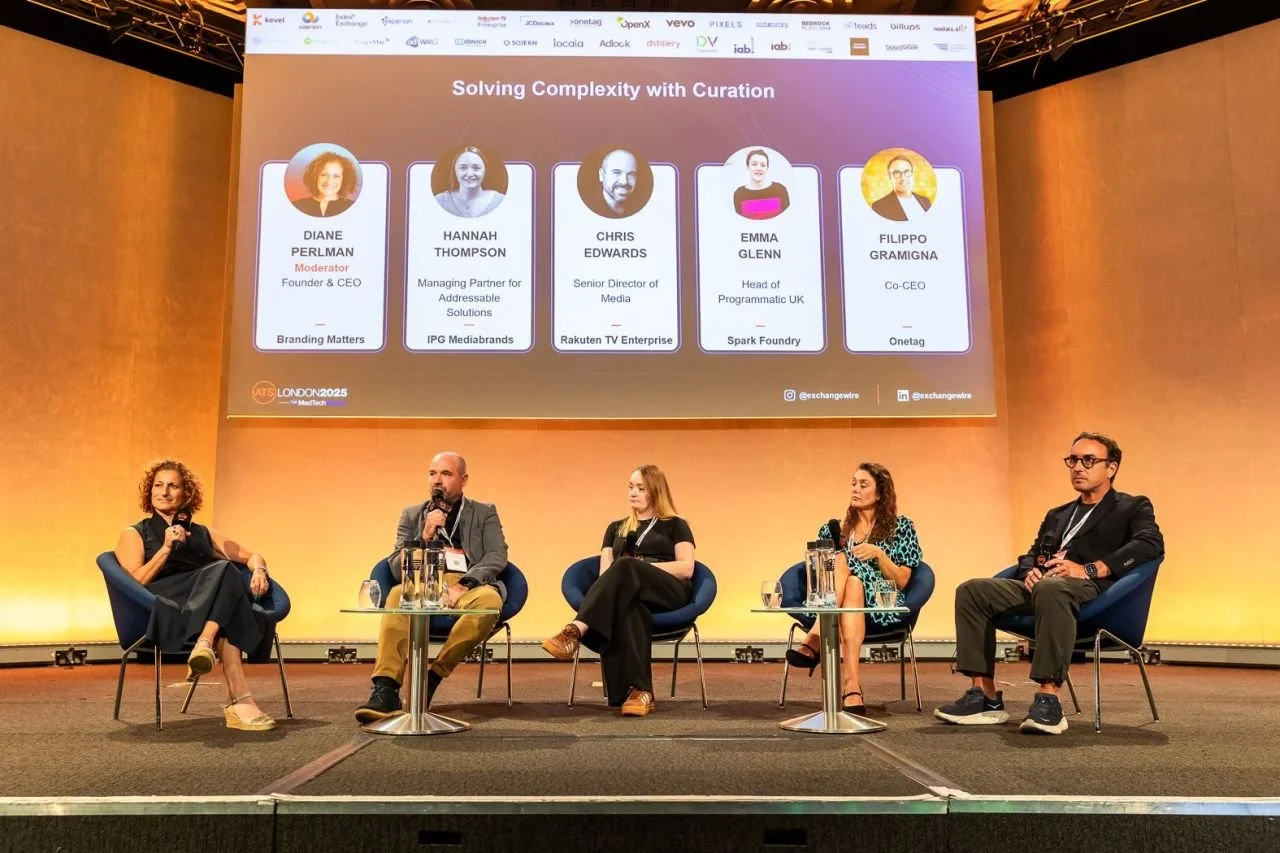 A panel discussion takes place on stage at a conference with five speakers seated in chairs. Behind them, a large screen displays the names and titles of the panelists, including moderator Diane Perlman and others from various media and tech companies. The stage has a warm, orange-toned background.