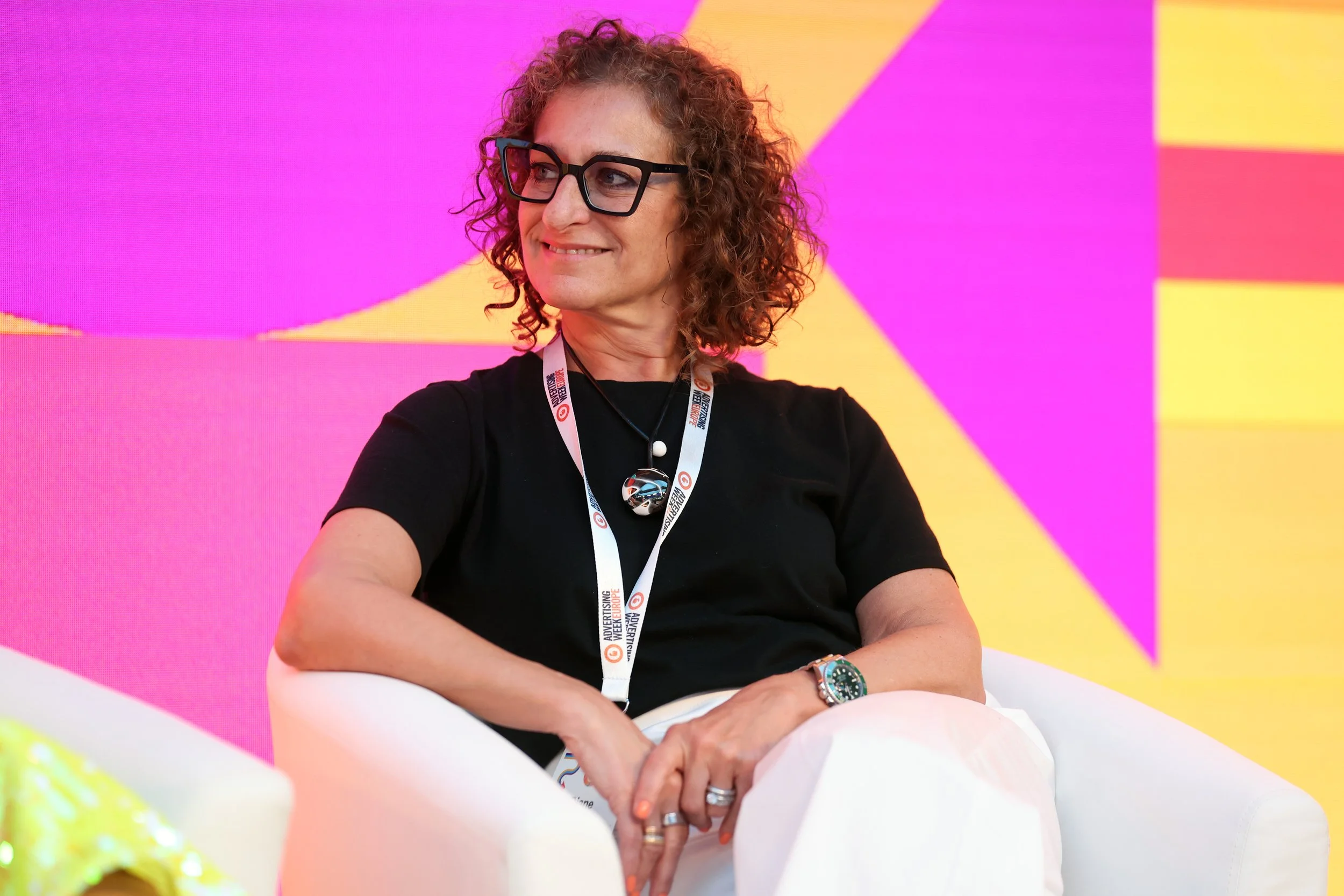 A woman with curly brown hair, wearing glasses and a black shirt, sitting with her arms crossed on her lap, smiling and looking to her right. She has a lanyard around her neck and a watch on her left wrist. The background features vibrant pink, yellow, and orange geometric shapes.