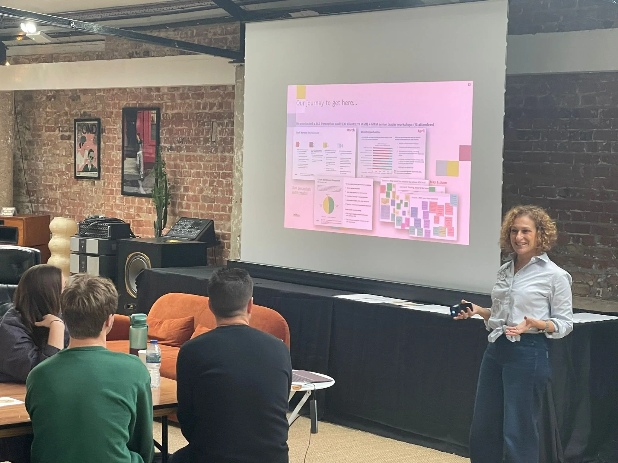 A woman is giving a presentation to a group of people in a room with exposed brick walls. There is a large pink poster with charts and graphs on the wall behind her. The audience is sitting at tables, some taking notes or listening attentively.