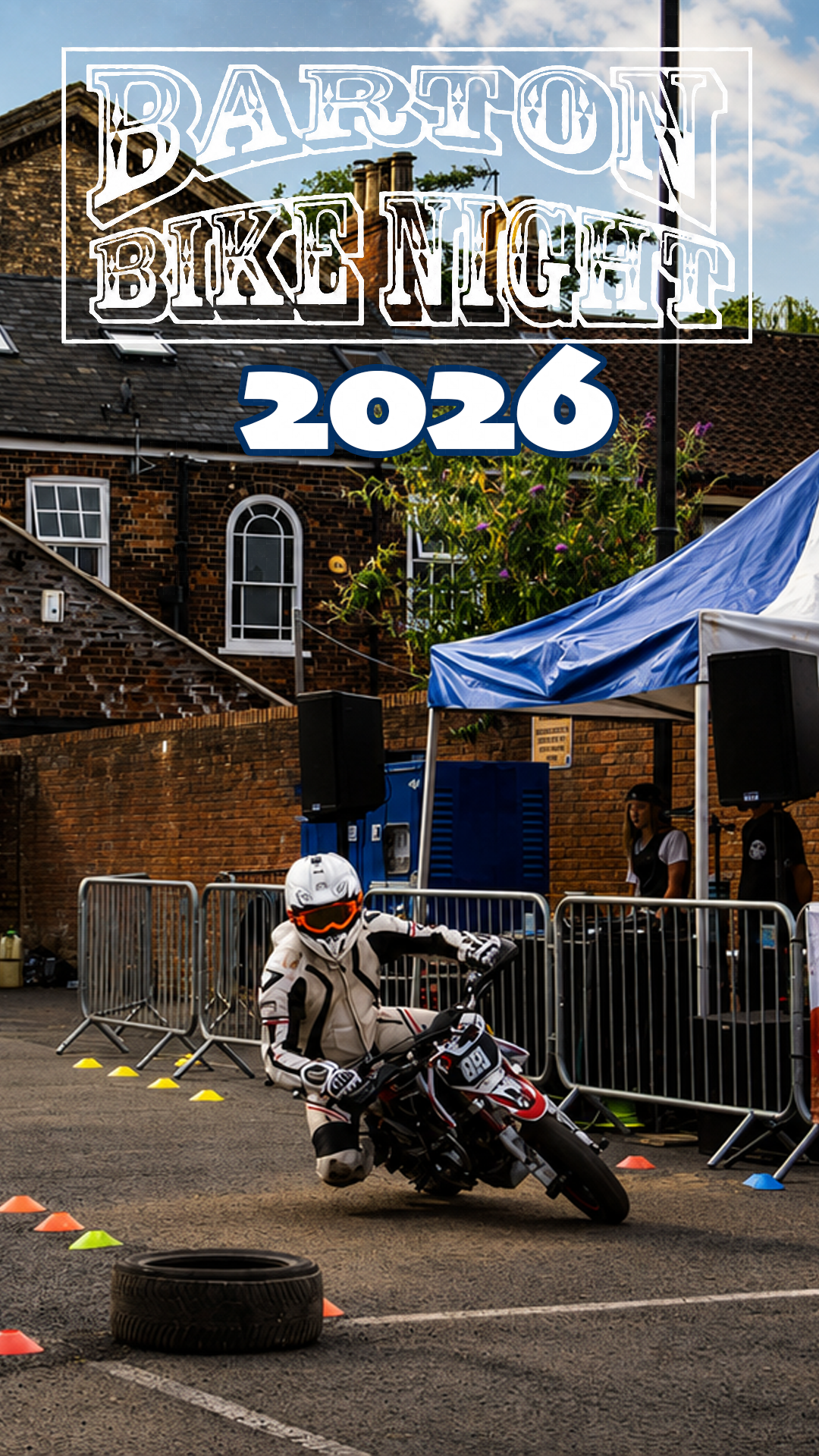 A motorcyclist in racing gear leaning into a turn on a motorcycle during a stunt event at Barton Bike Night 2026, with a brick building, tents, speakers, and spectators in the background.
