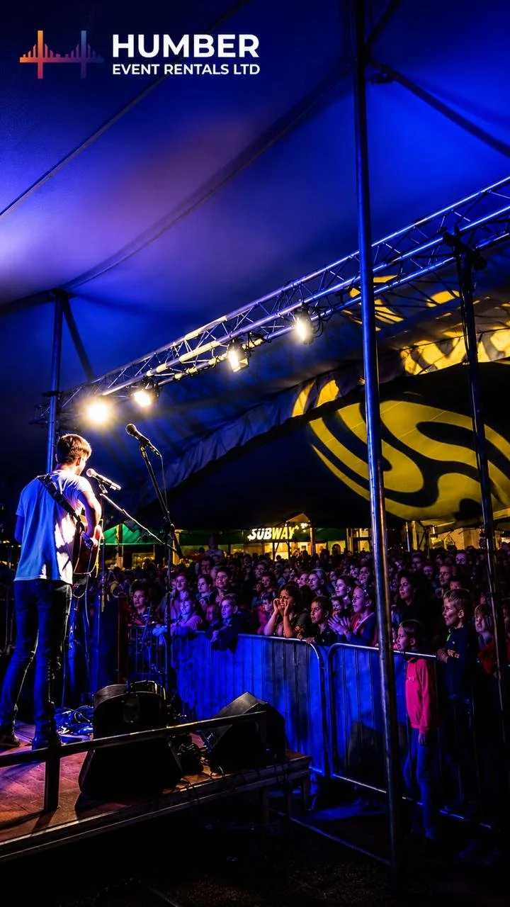 A musician performing on a stage under a canopy at night, with an audience of children and teens watching, illuminated by stage lights, and a Subway restaurant visible in the background.