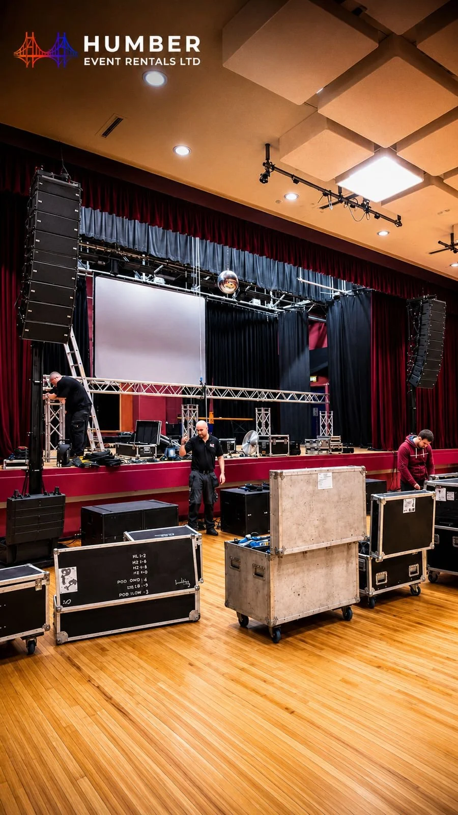 Stage setup with sound and lighting equipment, crew members preparing for an event at Humber Event Rentals, including large speakers, a projection screen, and various boxes filled with gear.
