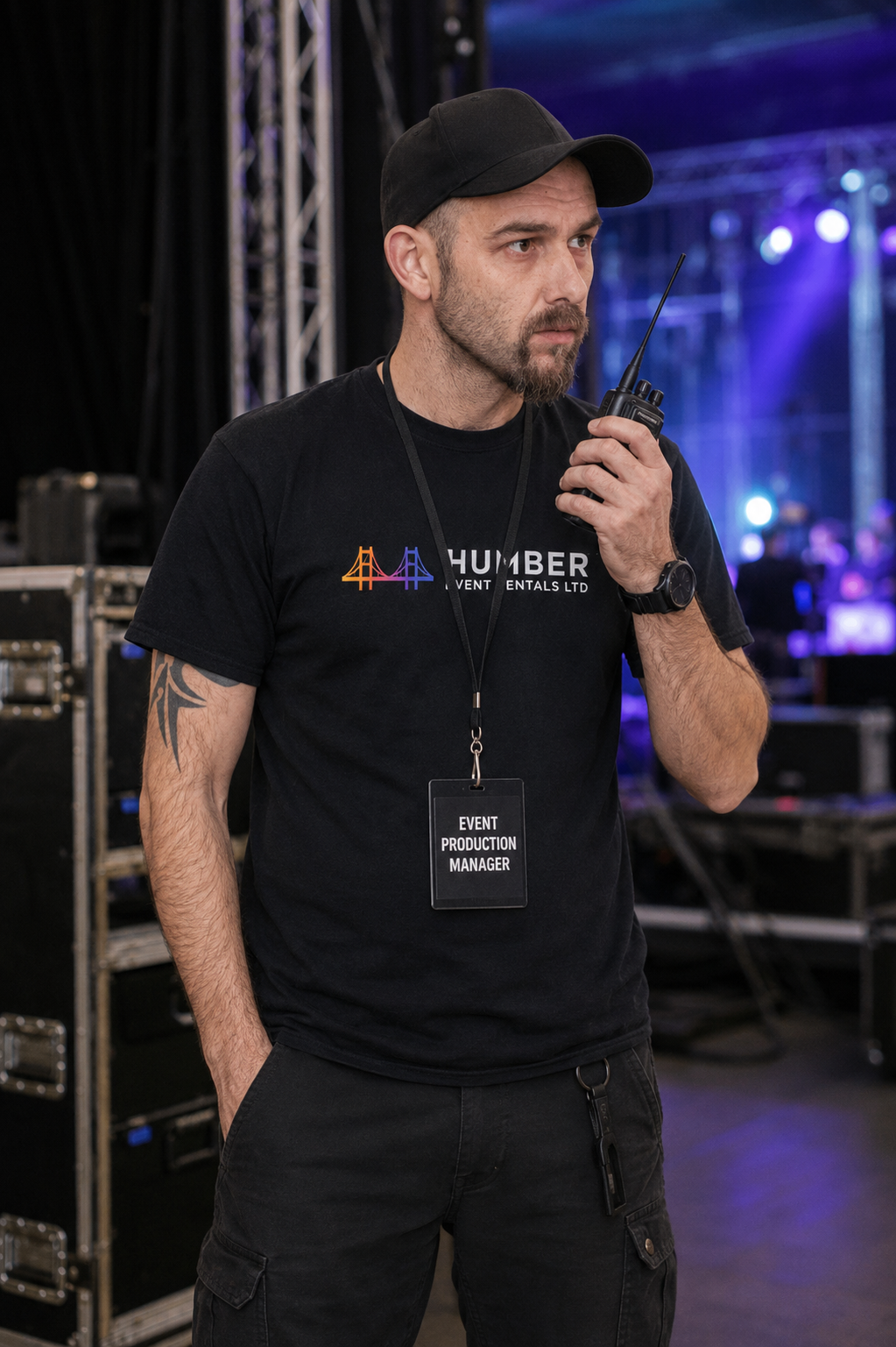Man in black T-shirt and cap holding walkie-talkie at event with purple lighting in background.