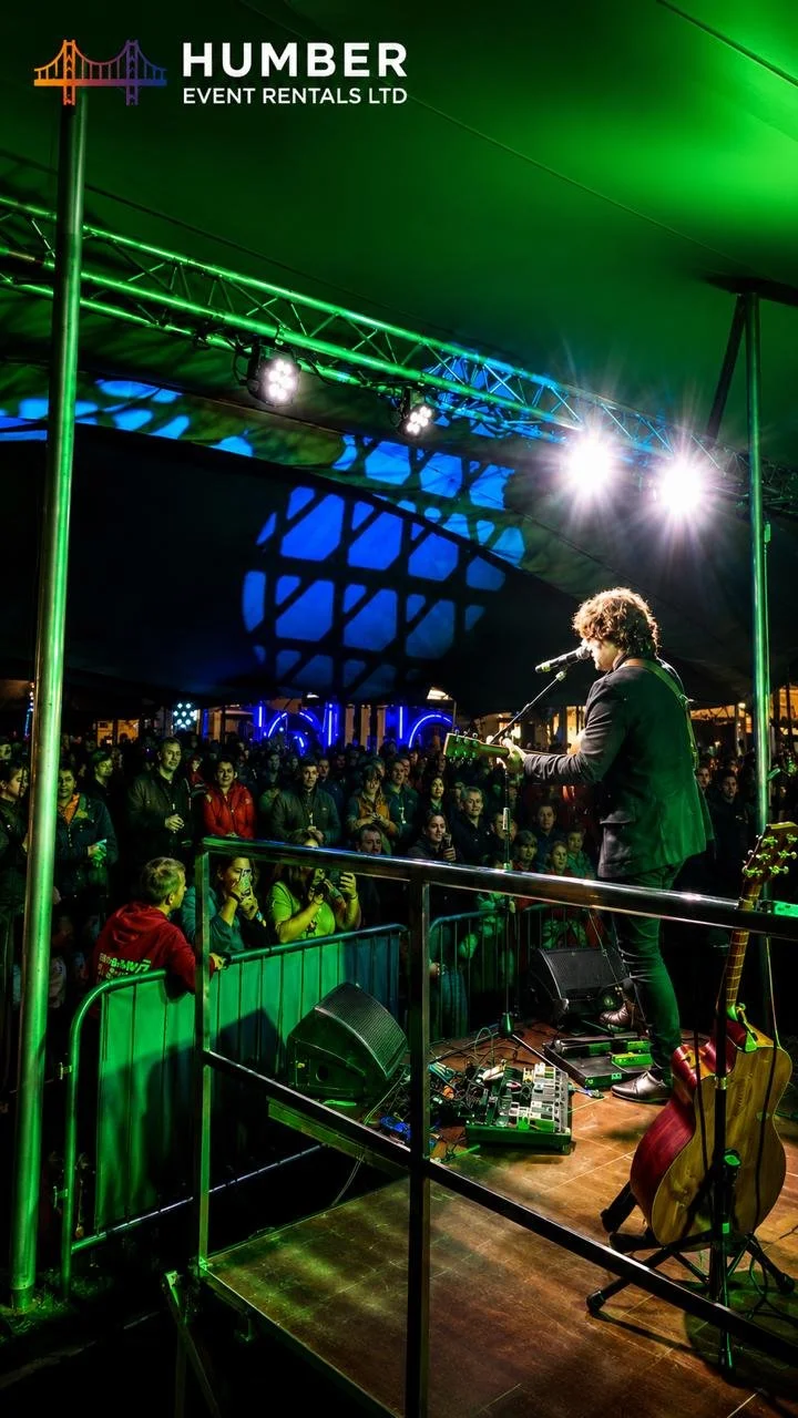 A musician performing on a stage at an event rental tent, with a crowd of people watching. The stage has colorful lighting, and the audience appears engaged, some taking photos. The tent has branding for Humber Event Rentals Ltd.