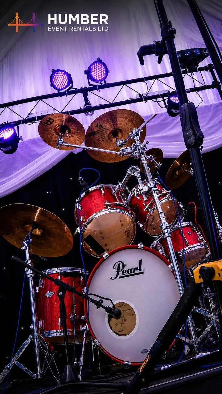 A red Pearl drum set on a stage with cymbals, drums, and microphone, under purple lighting with a Humber Event Rentals banner in the background.