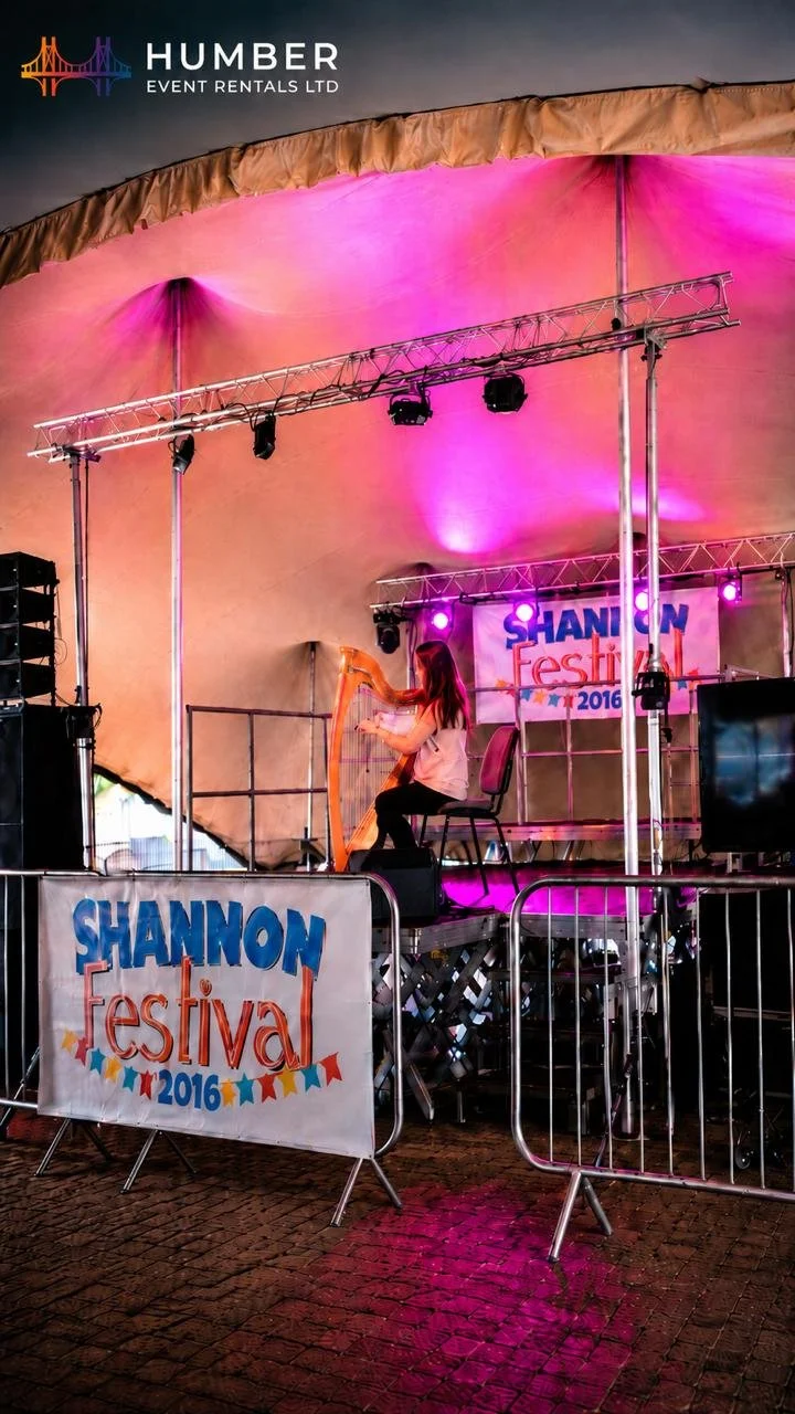 A young woman playing a harp on a stage at the Shannon Festival 2016, with pink and purple lighting, inside a large tent, with festival banners, speaker equipment, and metal barriers in front.