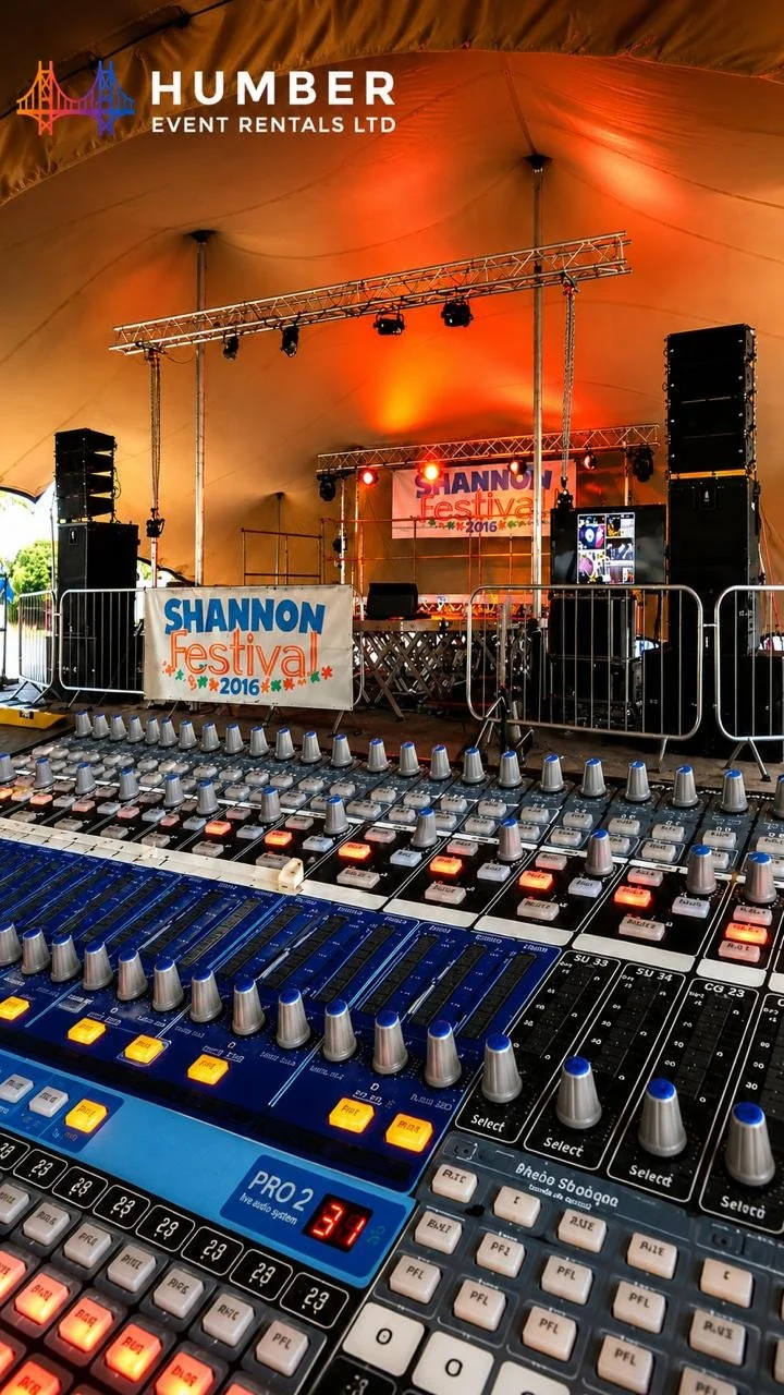 A sound mixing board in the foreground with knobs and sliders, and a stage with lighting and speakers in the background at the Shannon Festival 2016 event tent.