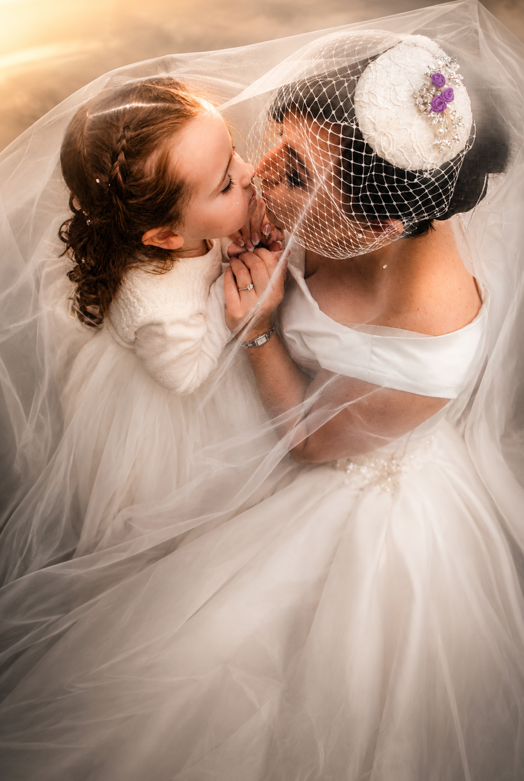A bride in a white wedding dress and veil with a flower bun, holding hands with a young girl dressed in a white dress, both leaning in for a kiss surrounded by soft, warm lighting.
