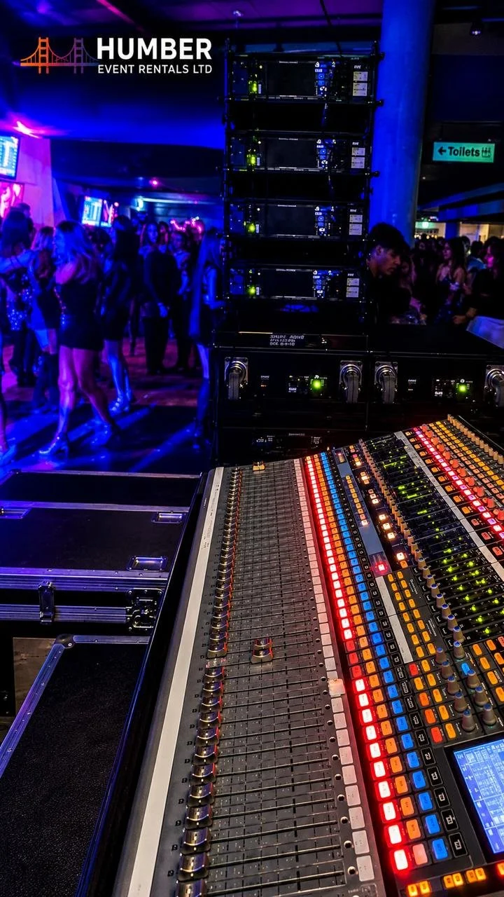 A sound mixing console in a nightclub or concert venue with a crowd of people dancing in the background, seen under blue and purple lighting.