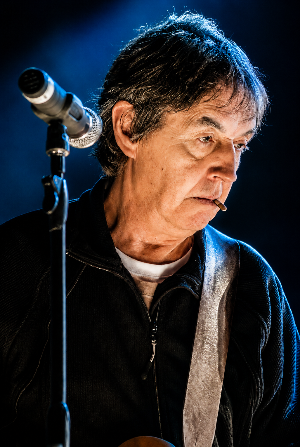 An older man with gray hair standing in front of a microphone, smoking a cigarette, and looking down.