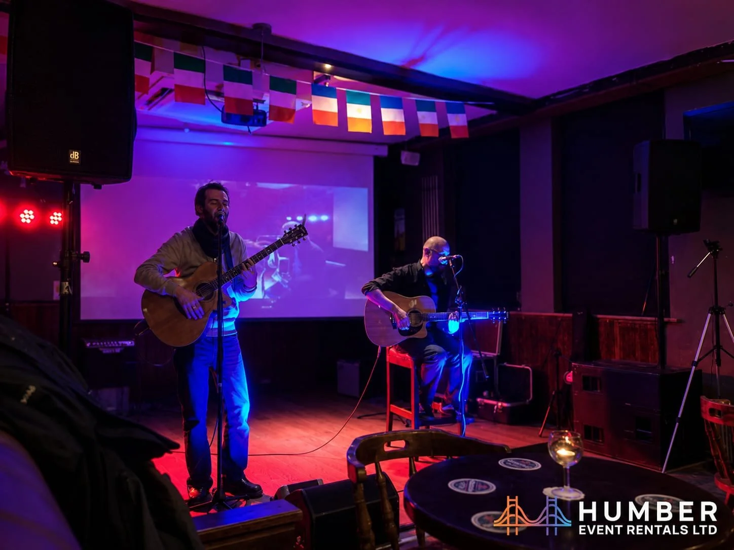 Two musicians playing acoustic guitars on a dimly lit stage with colorful lighting and a large screen displaying a live video feed in the background. There are flags hanging from the ceiling and a table with a glass of wine in the foreground, with a 