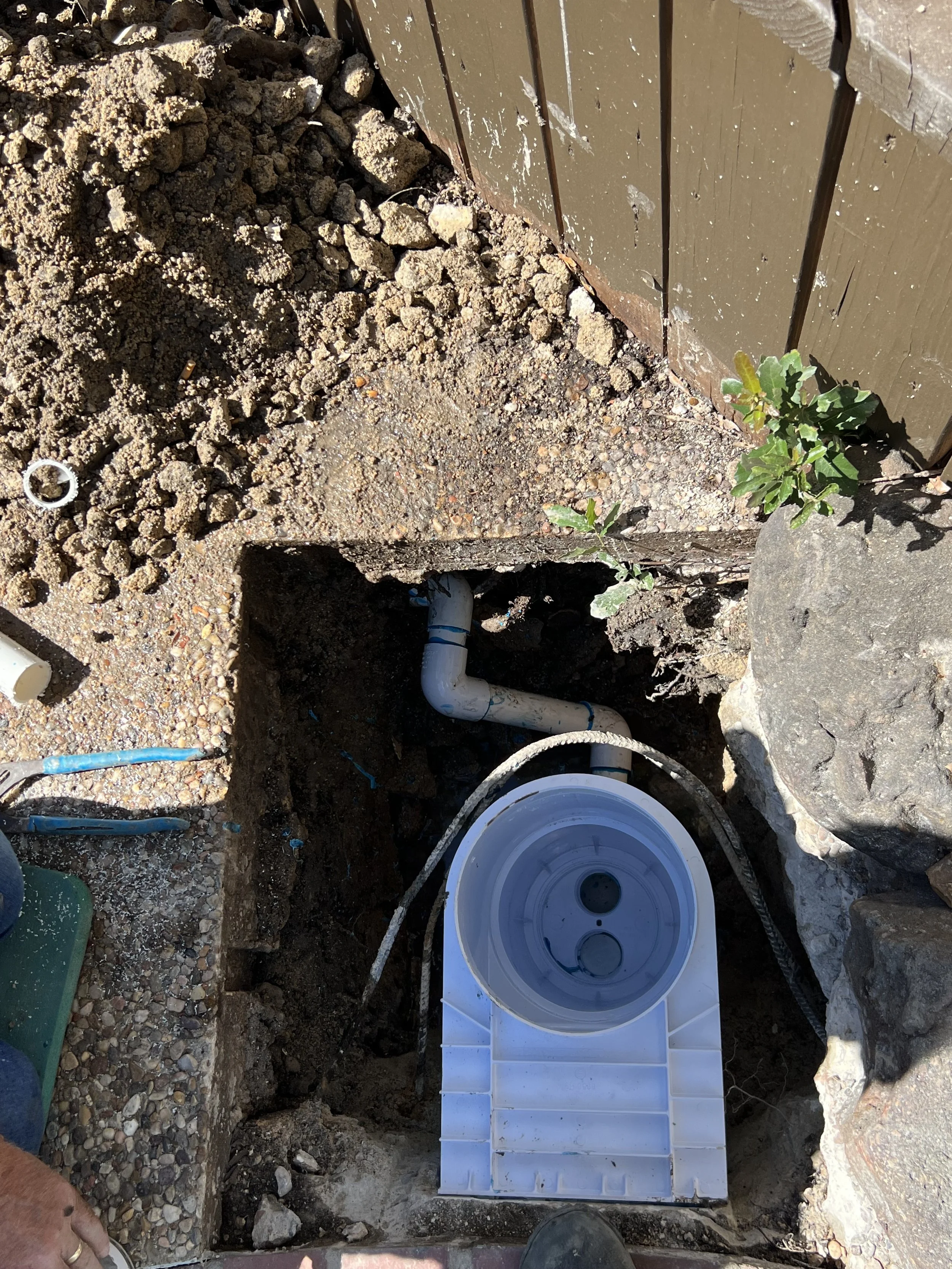A construction site showing a partially dug trench with a white plastic pipe installed at the bottom, surrounded by dirt, rocks, and some green plants near the edge.