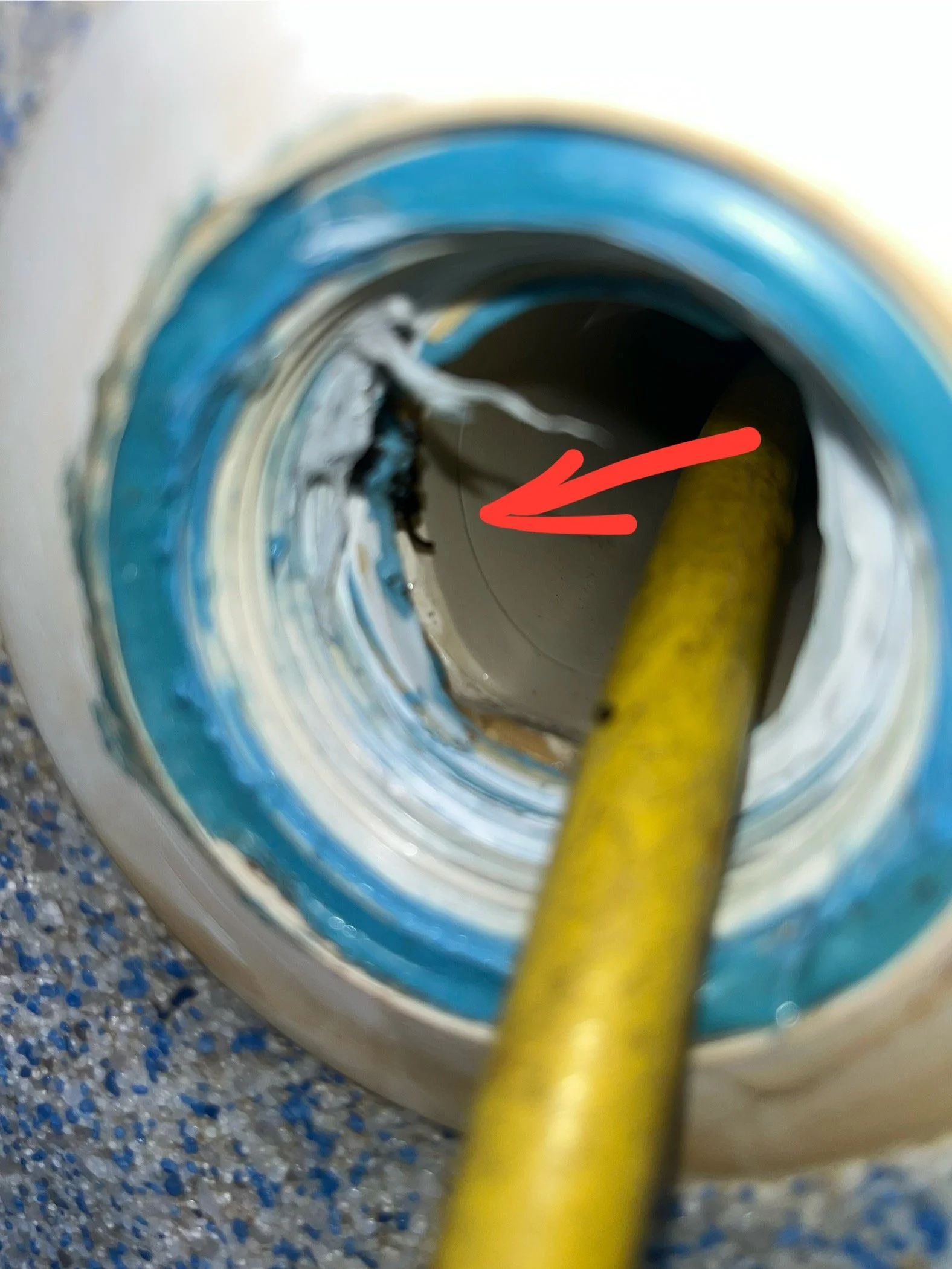 Close-up view of a gray pipe with blue sealing tape around it, with a yellow wire or cable inside the pipe, pointing towards the interior. The background shows a speckled gray and blue surface.