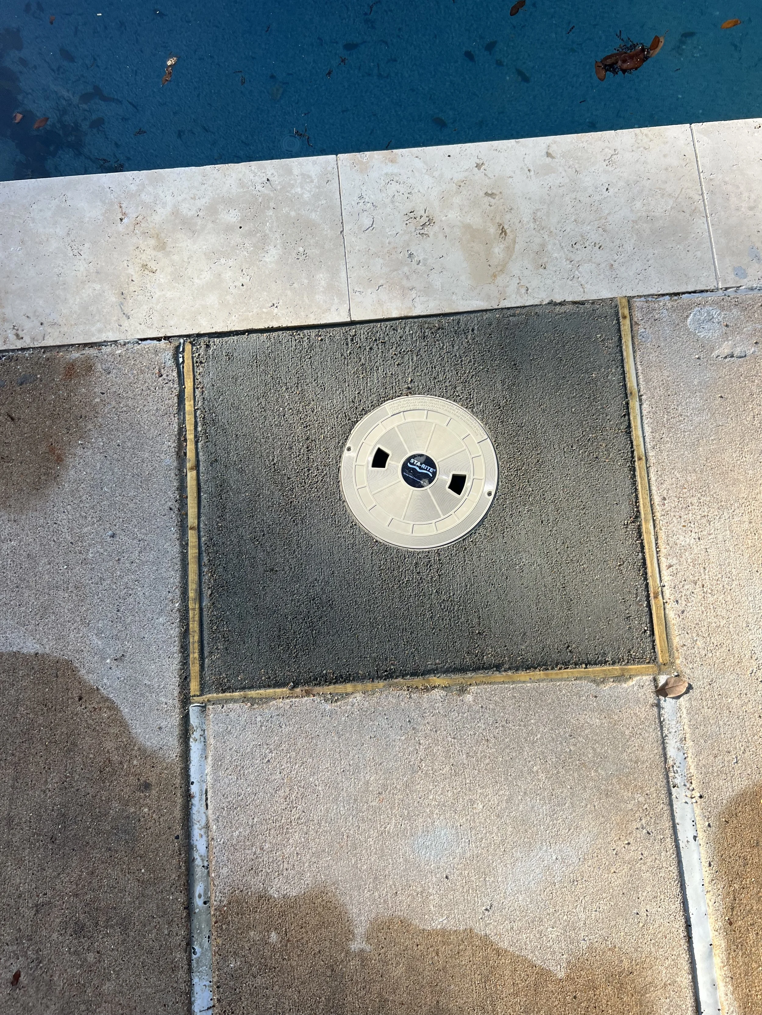 A round drain cover on a concrete pavement next to a swimming pool, with a corner of the pool visible at the top of the image.