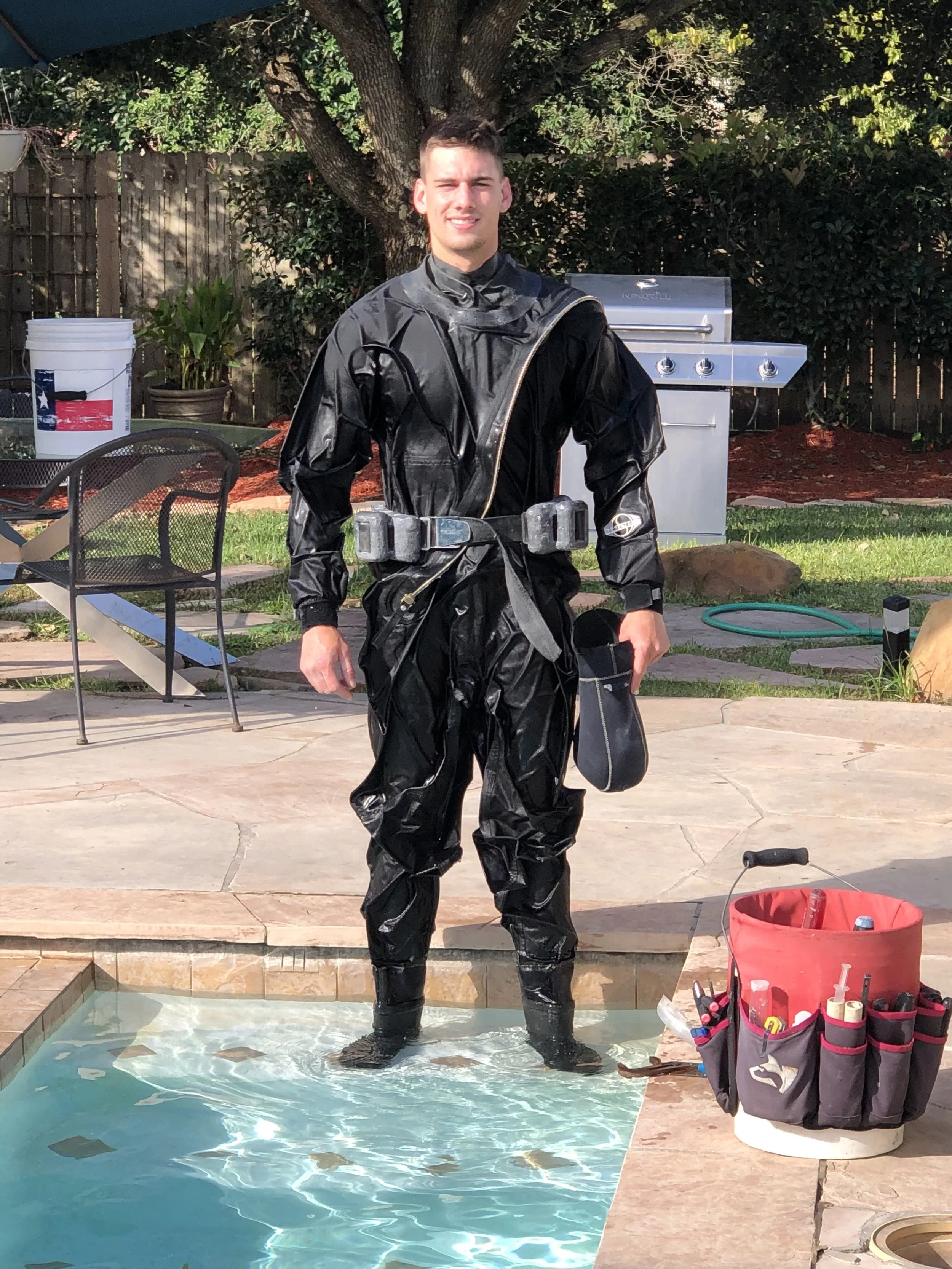 A young man standing in a small pool of water outdoors, wearing a black wetsuit. A large tree and a barbecue grill are in the background. There is a red bucket with tools and a tool bag nearby.
