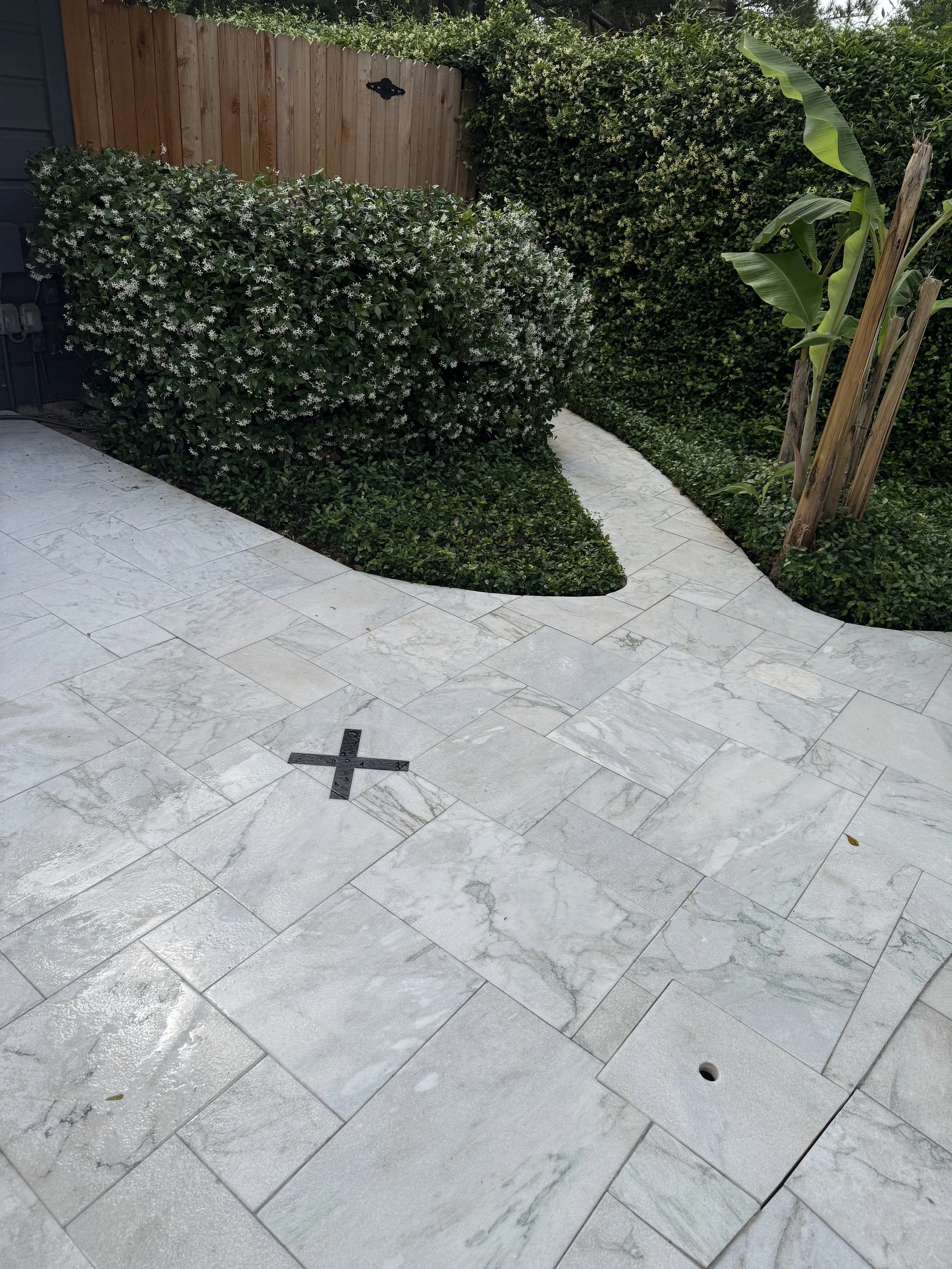 A backyard with a curved light-colored tiled patio, a bush with small white flowers, a wooden fence, and some banana plants supported by wooden stakes.