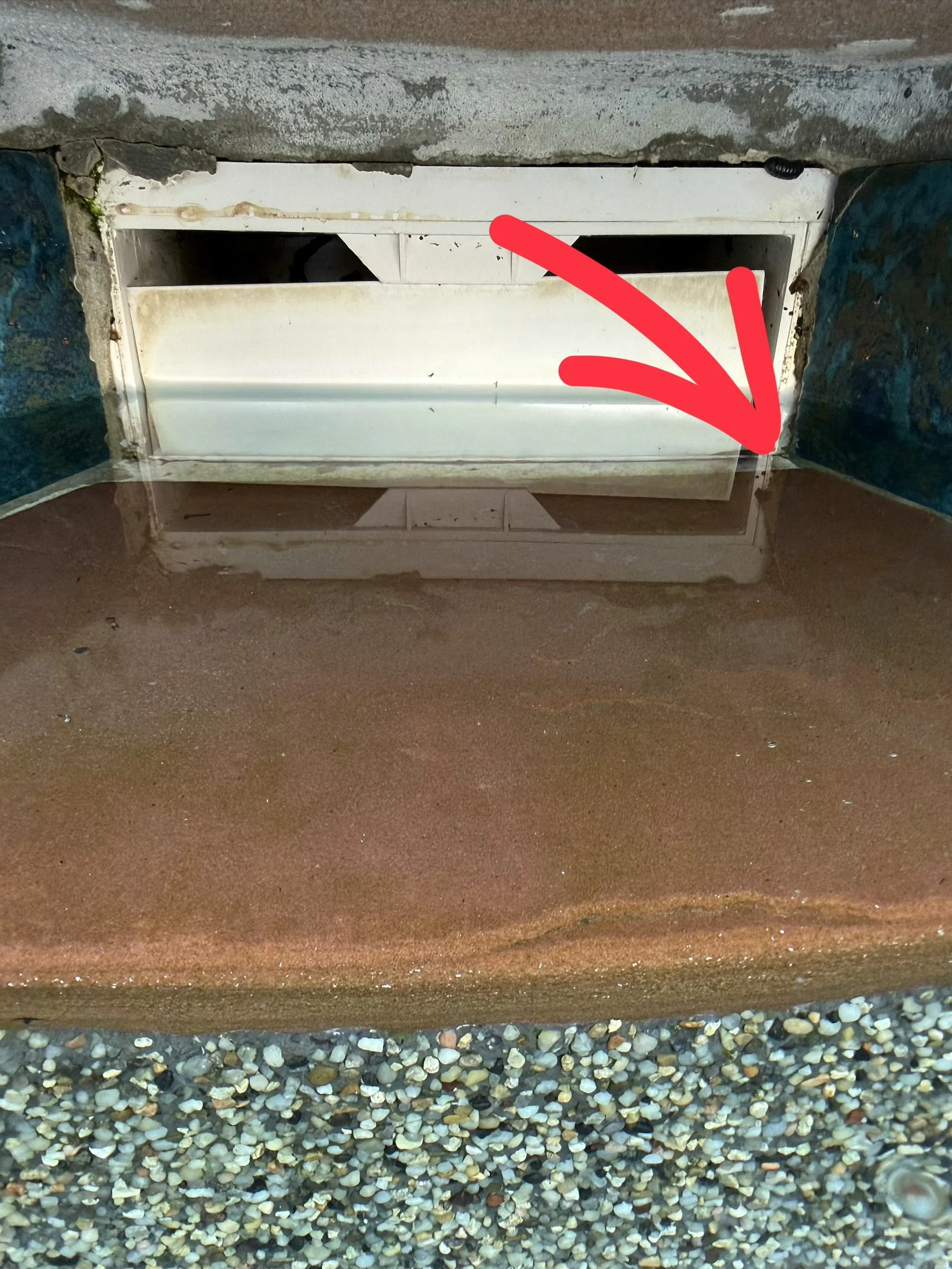Close-up of a vent or grille beneath a wooden or concrete surface, with a red arrow pointing at the bottom right corner of the vent. The vent is surrounded by blue-green tiles and small gravel or pebbles beneath.