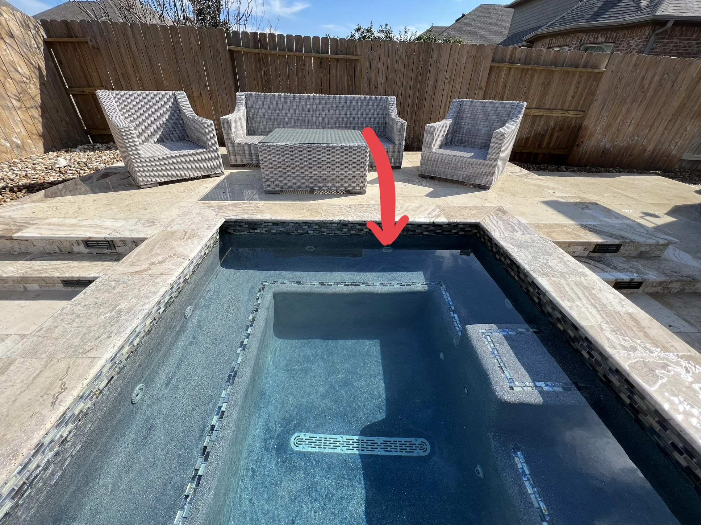 Patio area with outdoor furniture including a sofa, two armchairs, and a coffee table, surrounded by a wooden privacy fence. There is a swimming pool in the foreground with a decorative tile border and steps leading into the water. The area is paved with light-colored stone tiles, and a chimney is visible in the background. A red arrow is pointing towards a drain or sensor on the edge of the pool.