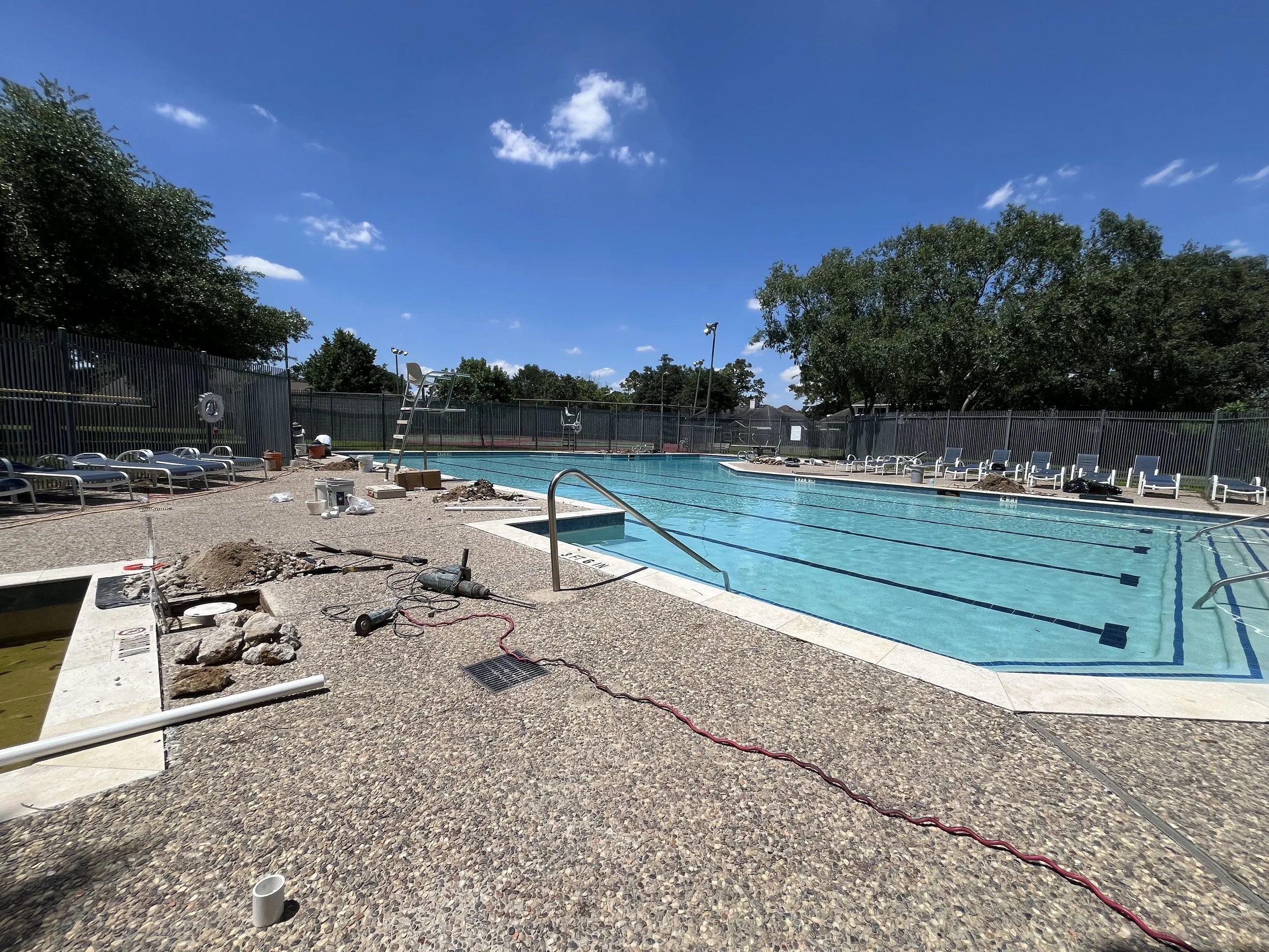 Public swimming pool under renovation with construction tools, pipes, and debris around the pool edge, surrounded by a black fence and trees.