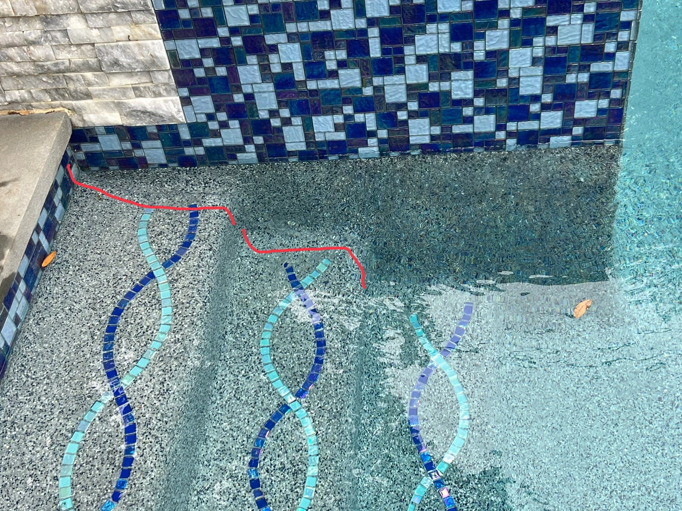 Close-up of a mosaic wall with small square tiles in various shades of blue and white, next to a textured concrete surface with curved lines made of small blue tiles, part of a pool. Water is visible in the lower right corner.