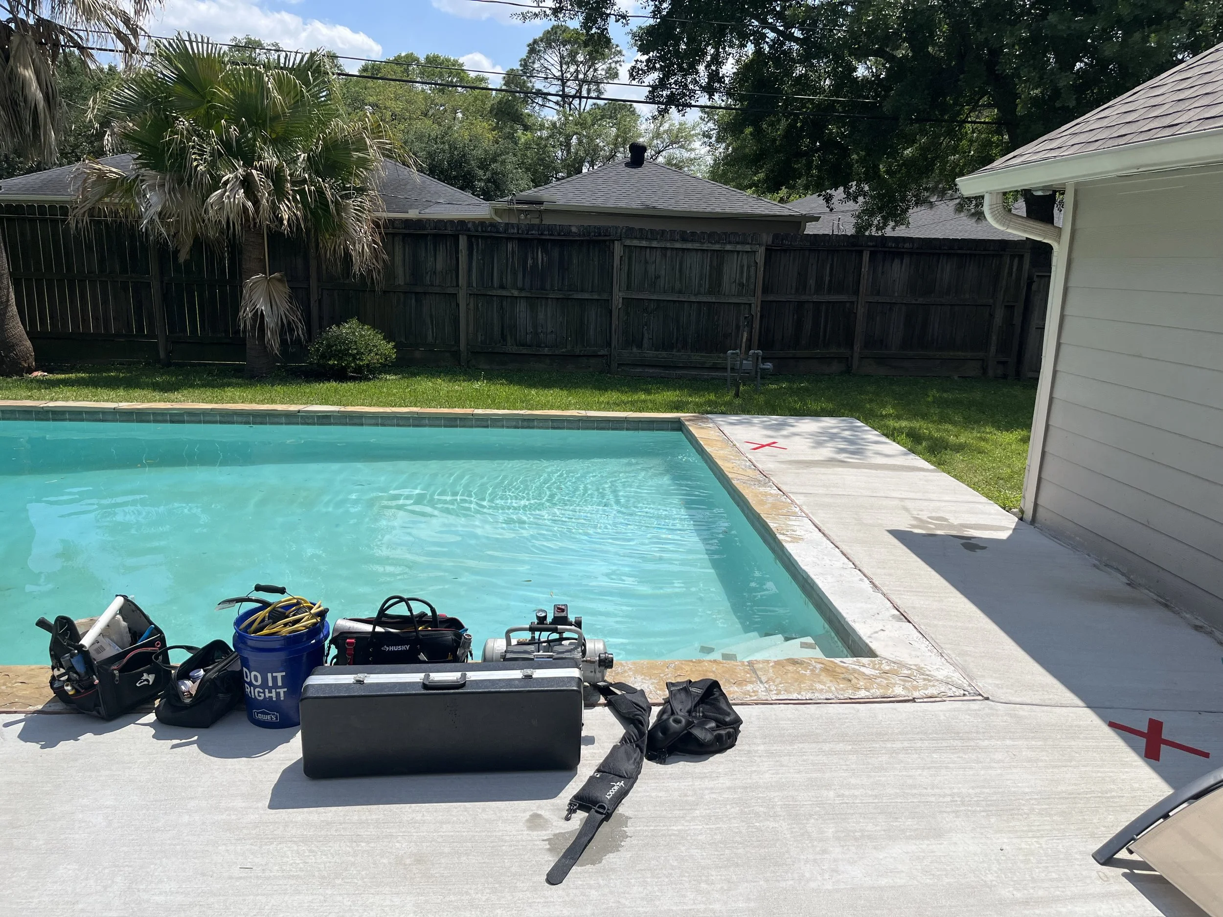 Backyard scene with a swimming pool under maintenance, tools and equipment for cleaning and repair are placed on the patio, and a wooden fence with trees and houses in the background.