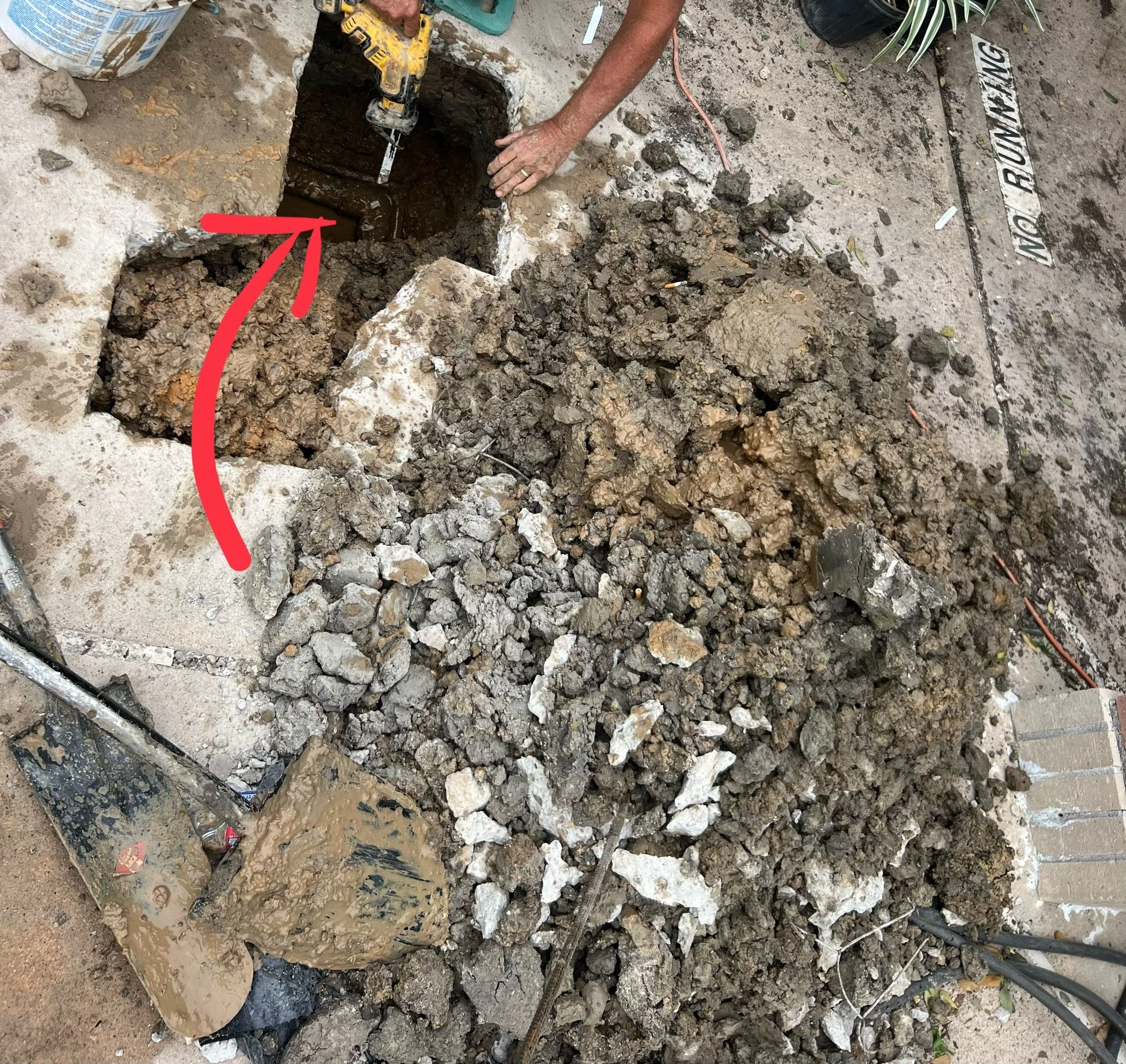 Person using a power tool to break up concrete in a hole in the ground, with rocks, debris, and tools scattered around.