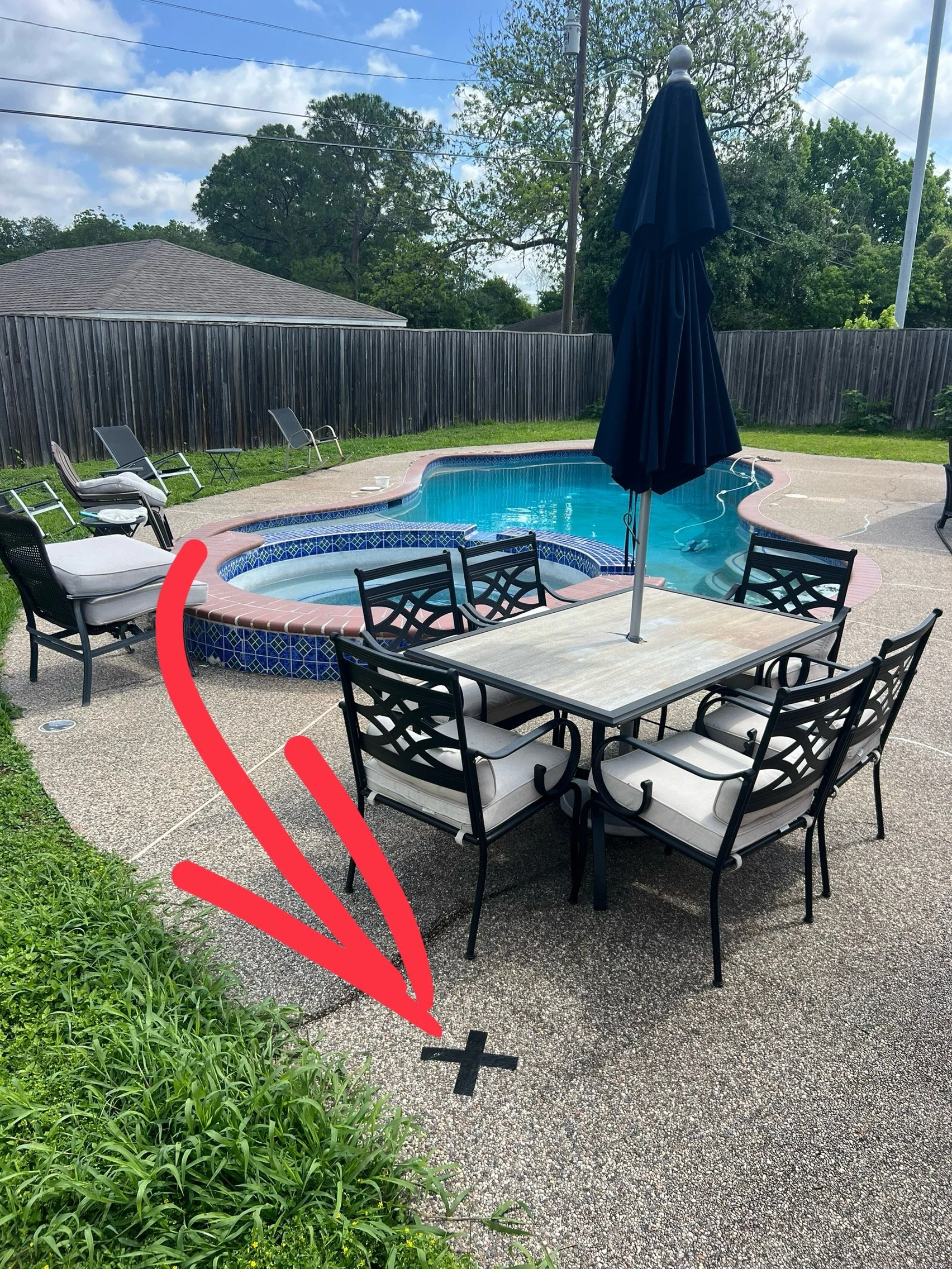 A backyard patio with a swimming pool, outdoor seating area with chairs and table, large umbrella, green grass, a wooden fence, trees, and a house roof in the background. There is also a marked black cross on the ground and an arrow pointing towards 
