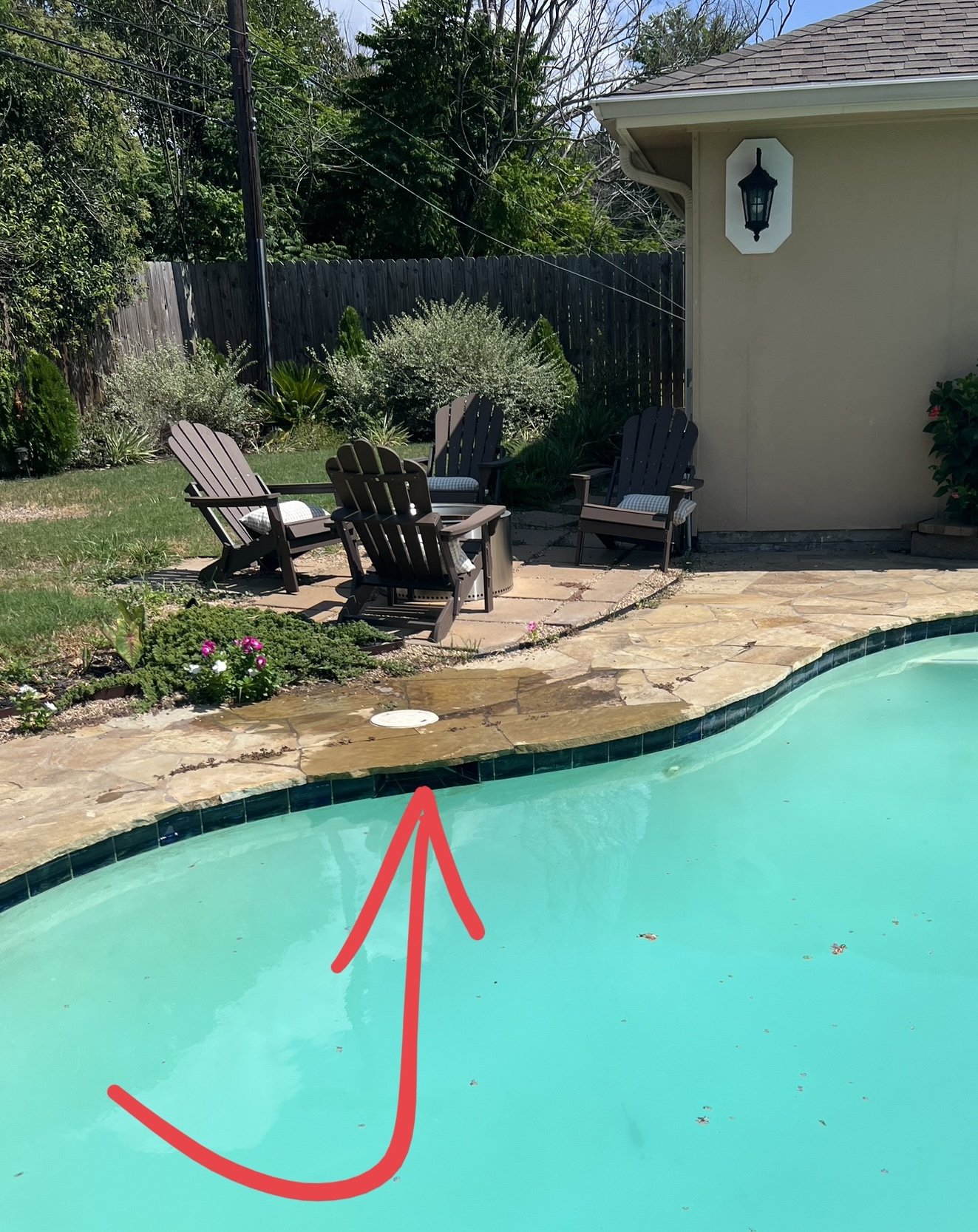 Backyard with a swimming pool, four Adirondack chairs around a fire pit, a garden with flowering plants, a fence, and part of a house with a lantern-style light fixture.