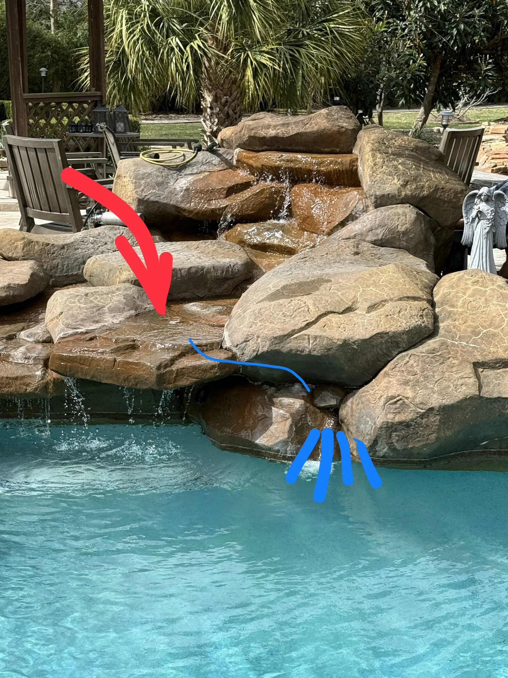 A backyard swimming pool with a rock waterfall feature, surrounded by patio furniture and landscaping, including a palm tree and other plants.